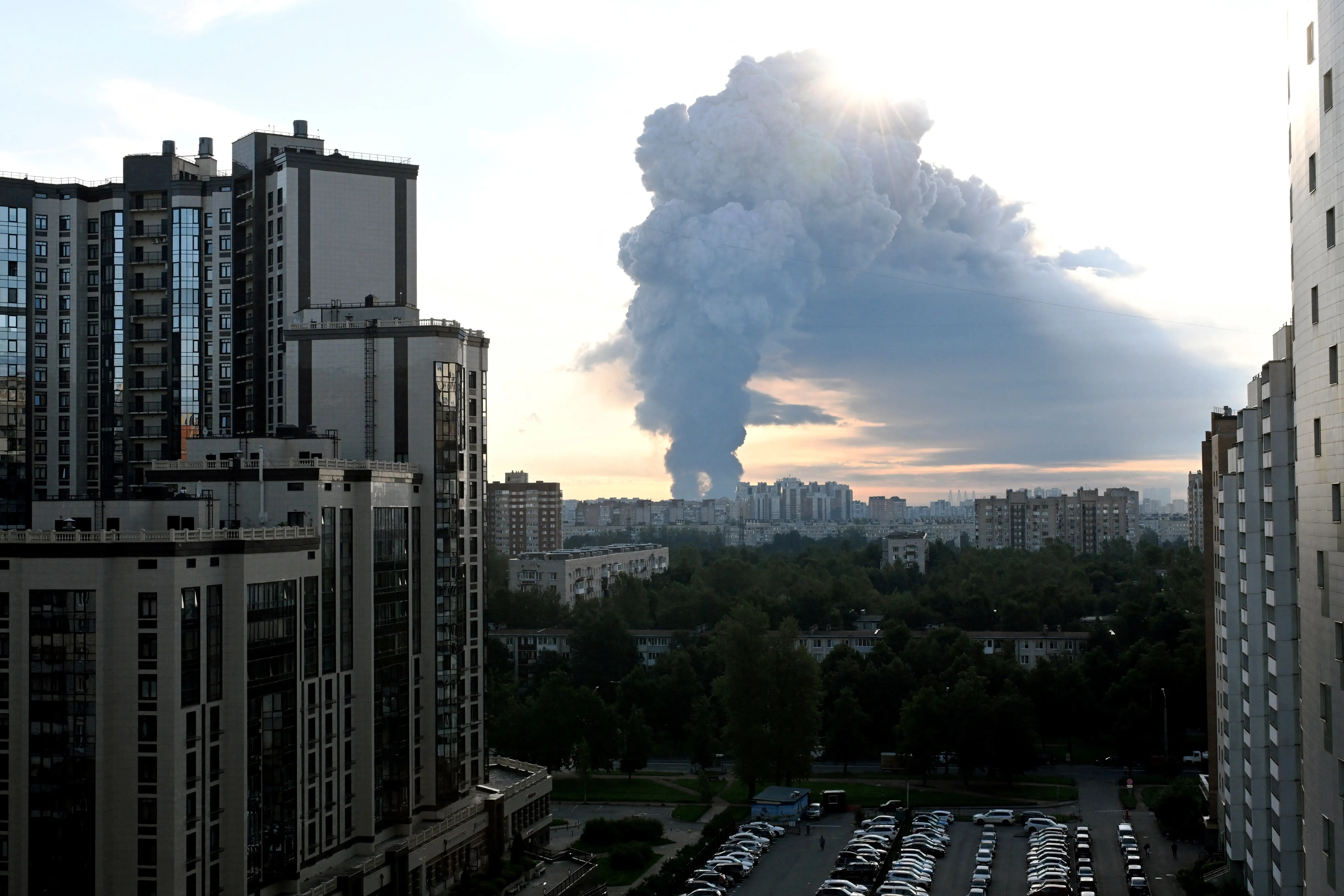 A large smoke plume rises beyond high-rise apartment buildings in an urban skyline at sunset.