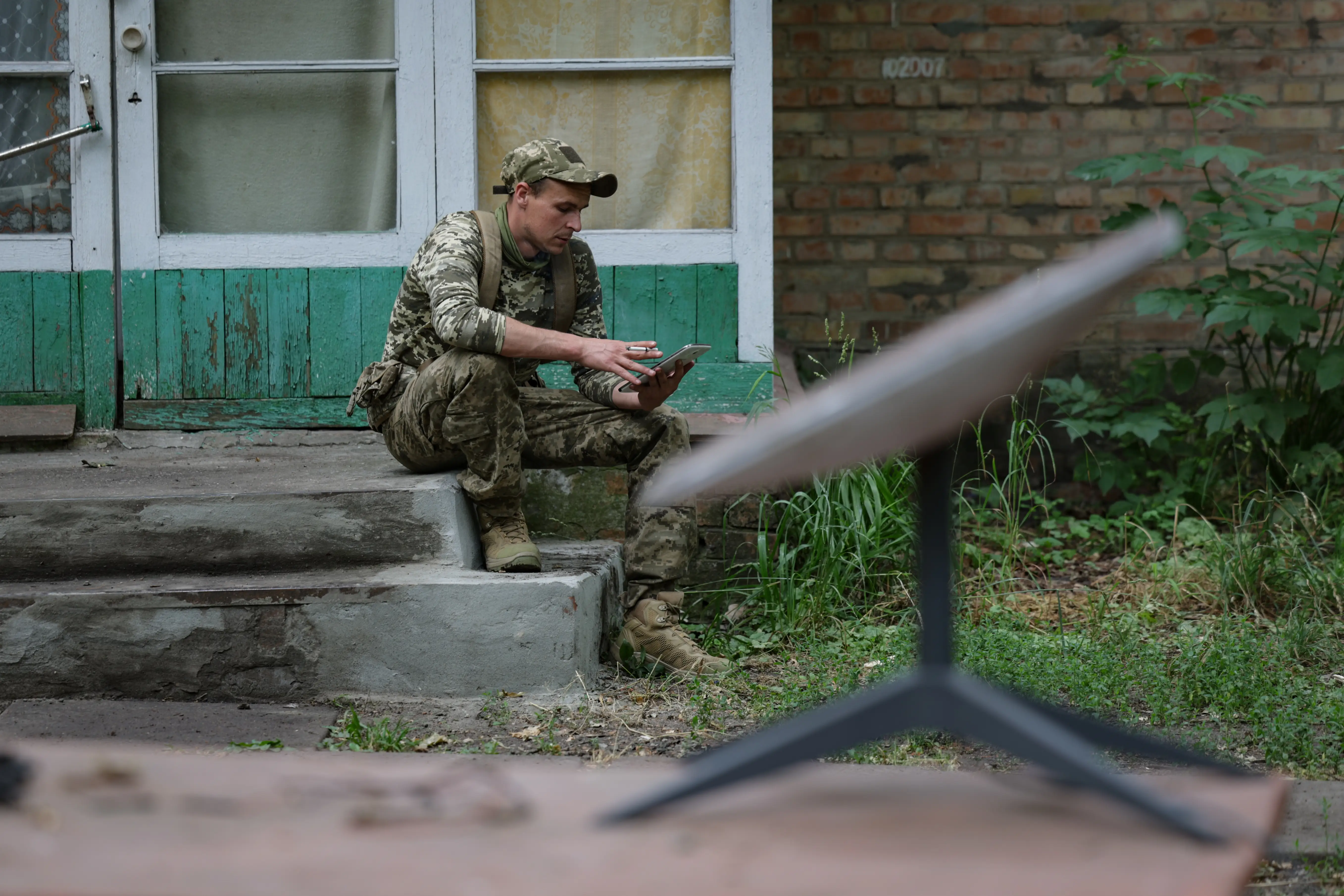 Person in camouflage uniform sits on concrete steps using a device near a satellite antenna outside a brick building.