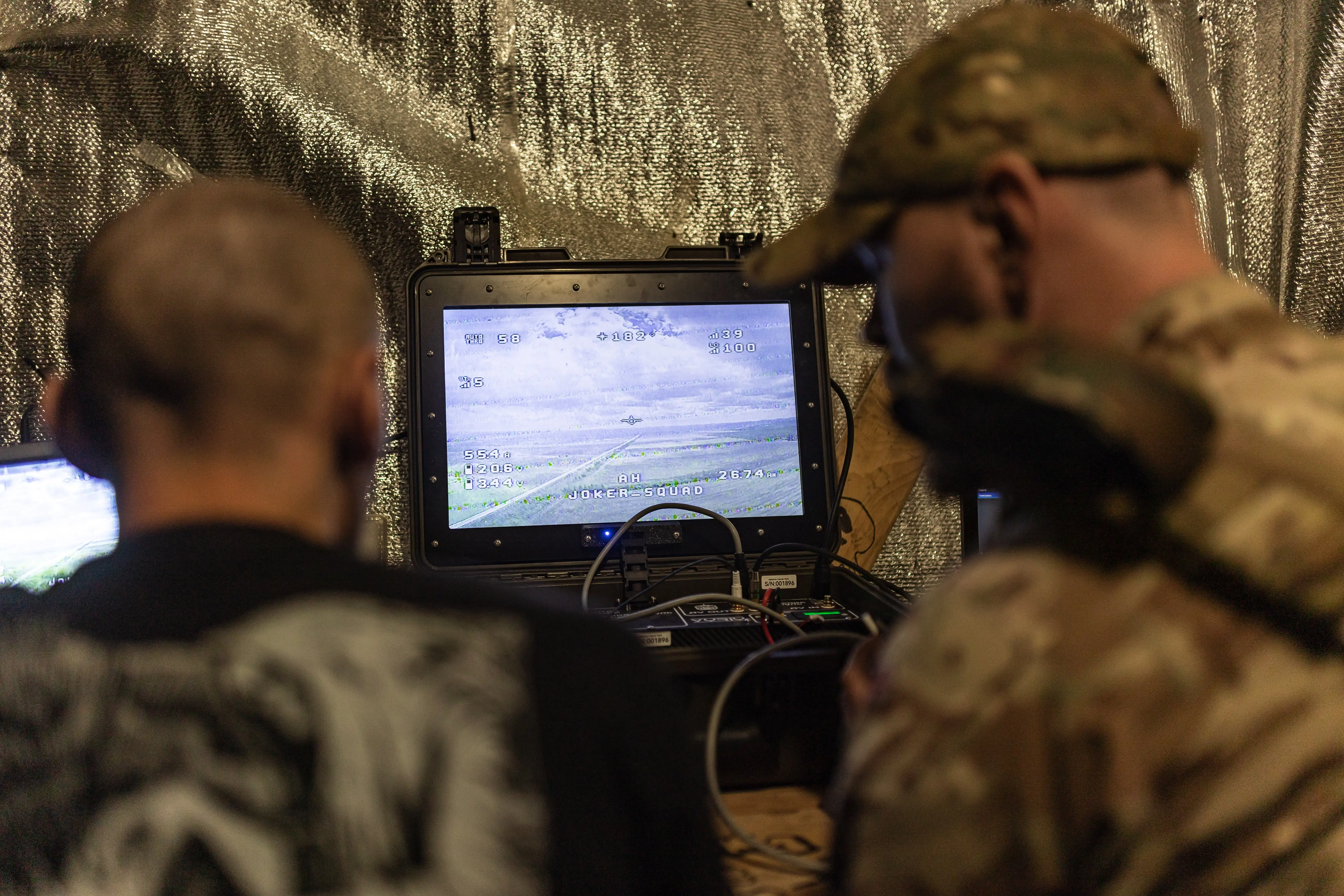 Two people view a monitor showing aerial video feeds inside a metallic-walled operations area.