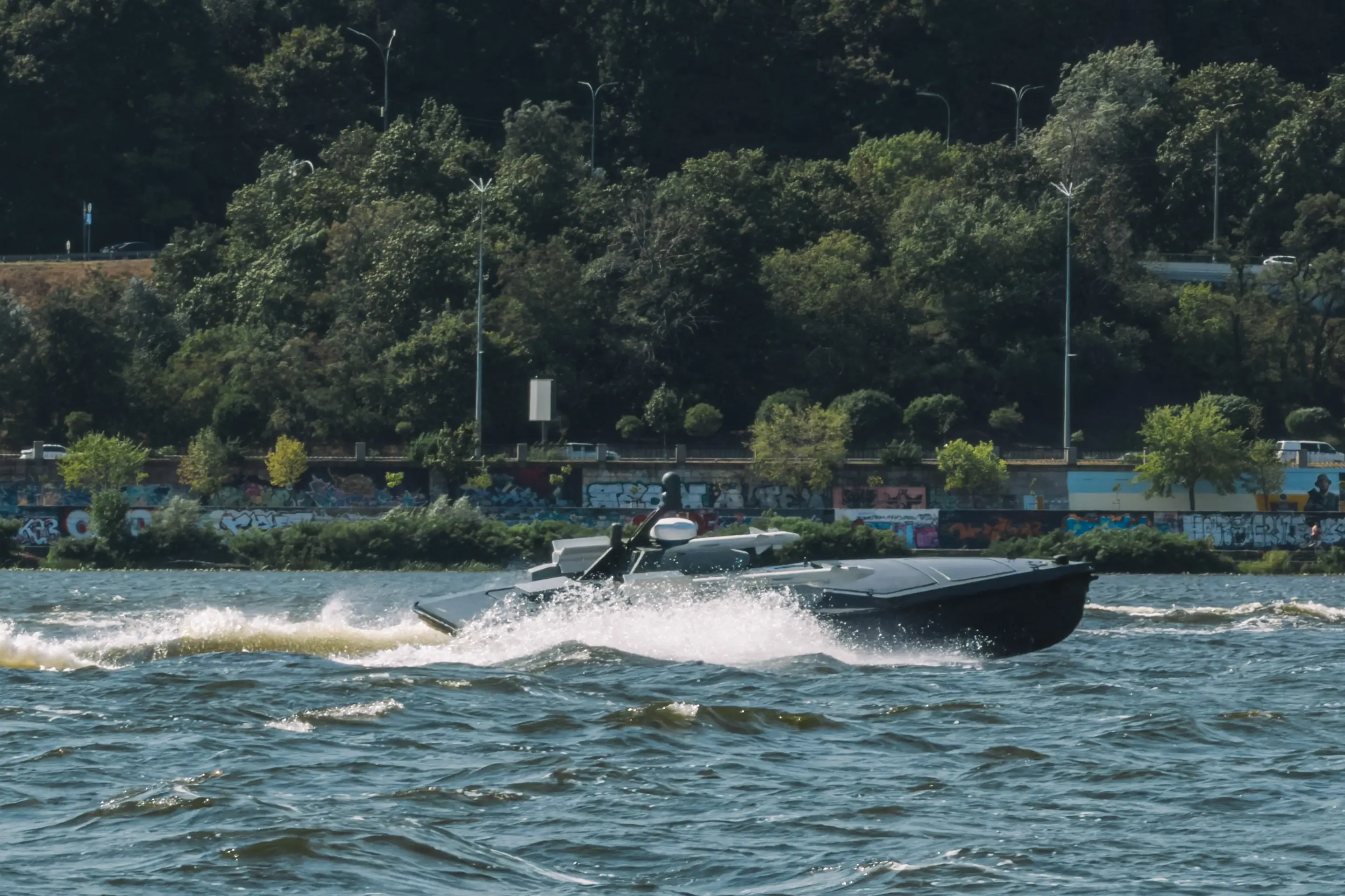 Black speedboat travels across choppy water near a wooded shoreline and graffiti-covered wall.
