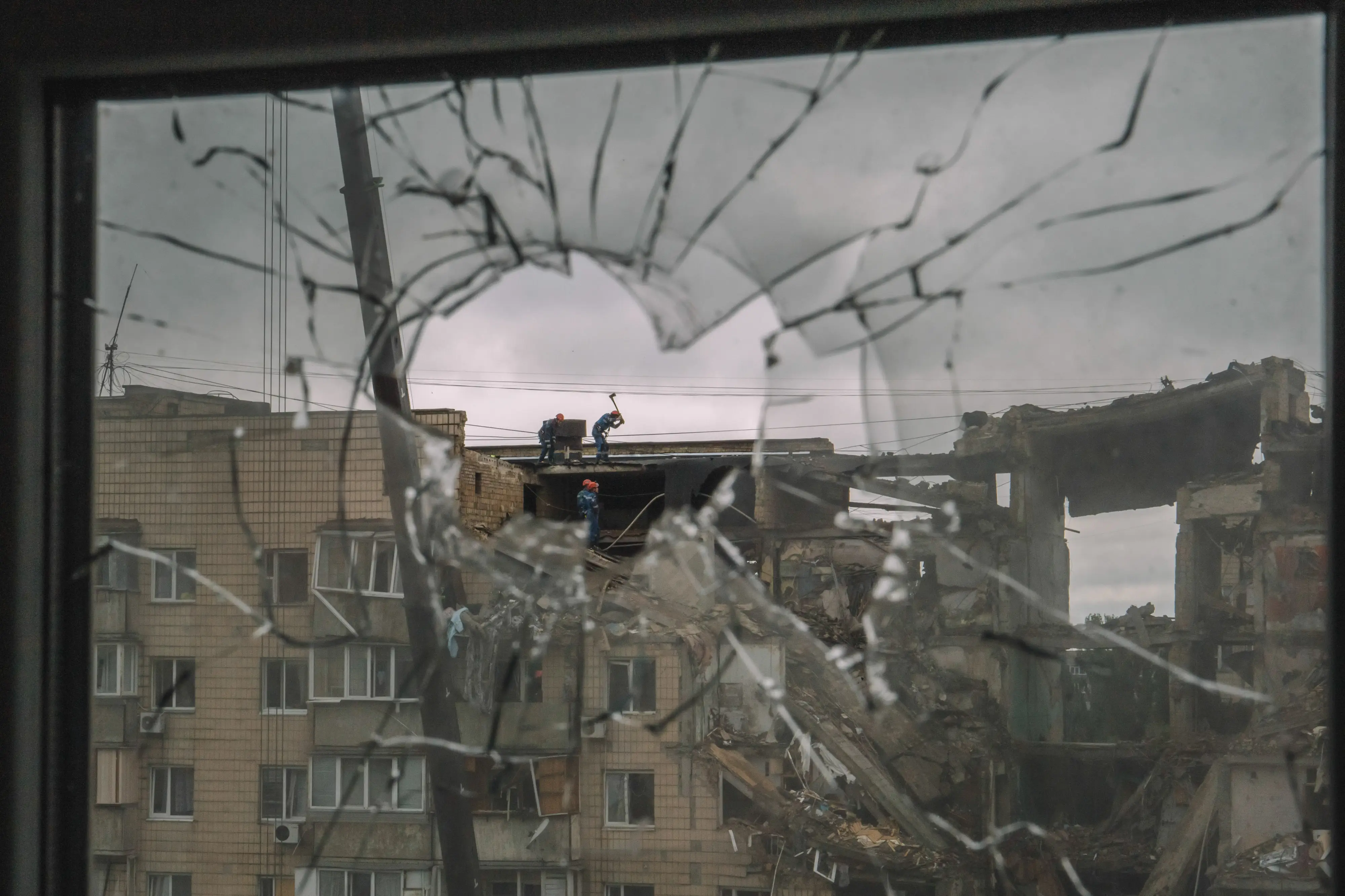 Shattered window glass frames a heavily damaged apartment building with several people standing on the roof.