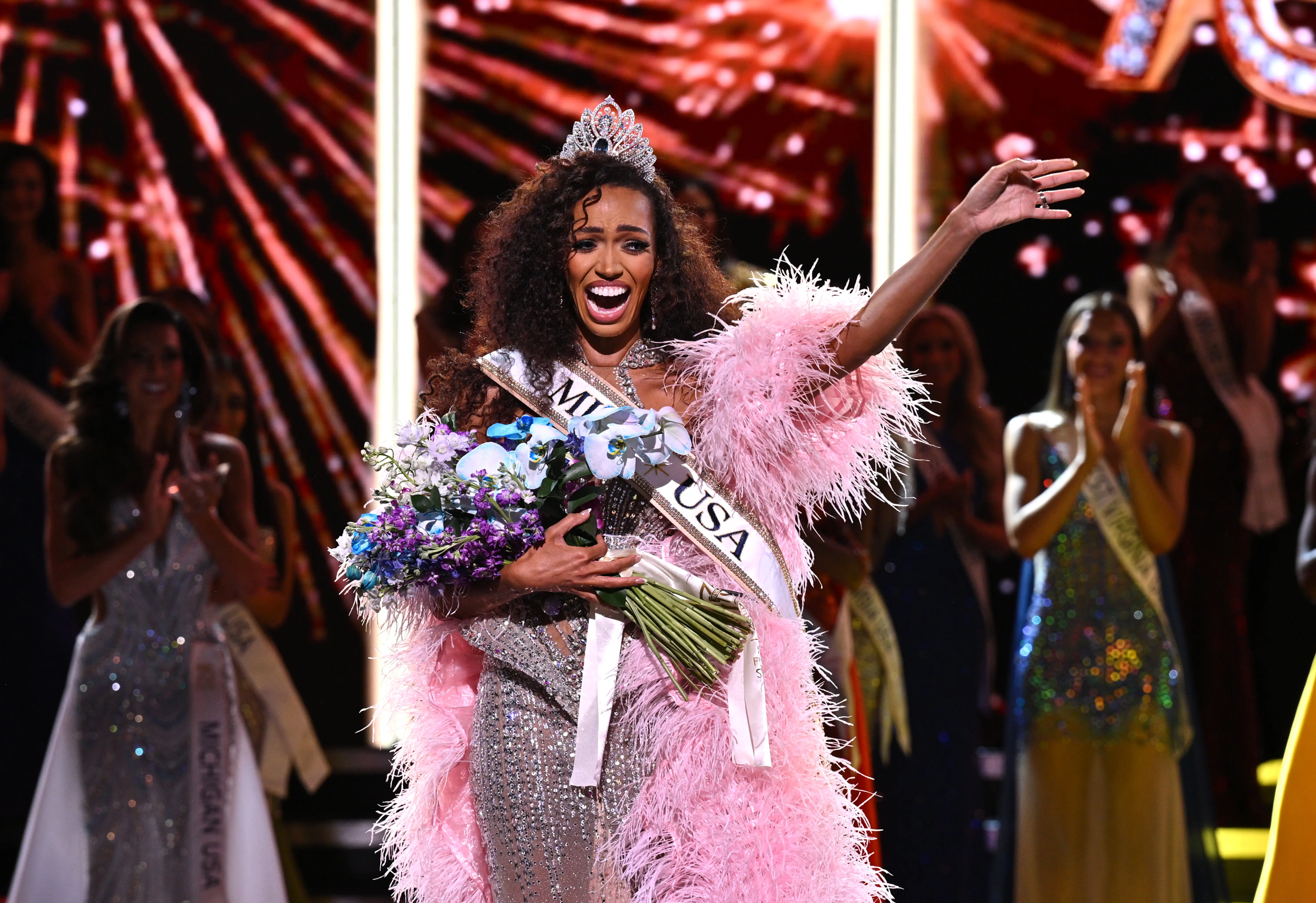 Crowned MISS USA pageant contestant waves while holding a bouquet on a brightly lit stage.