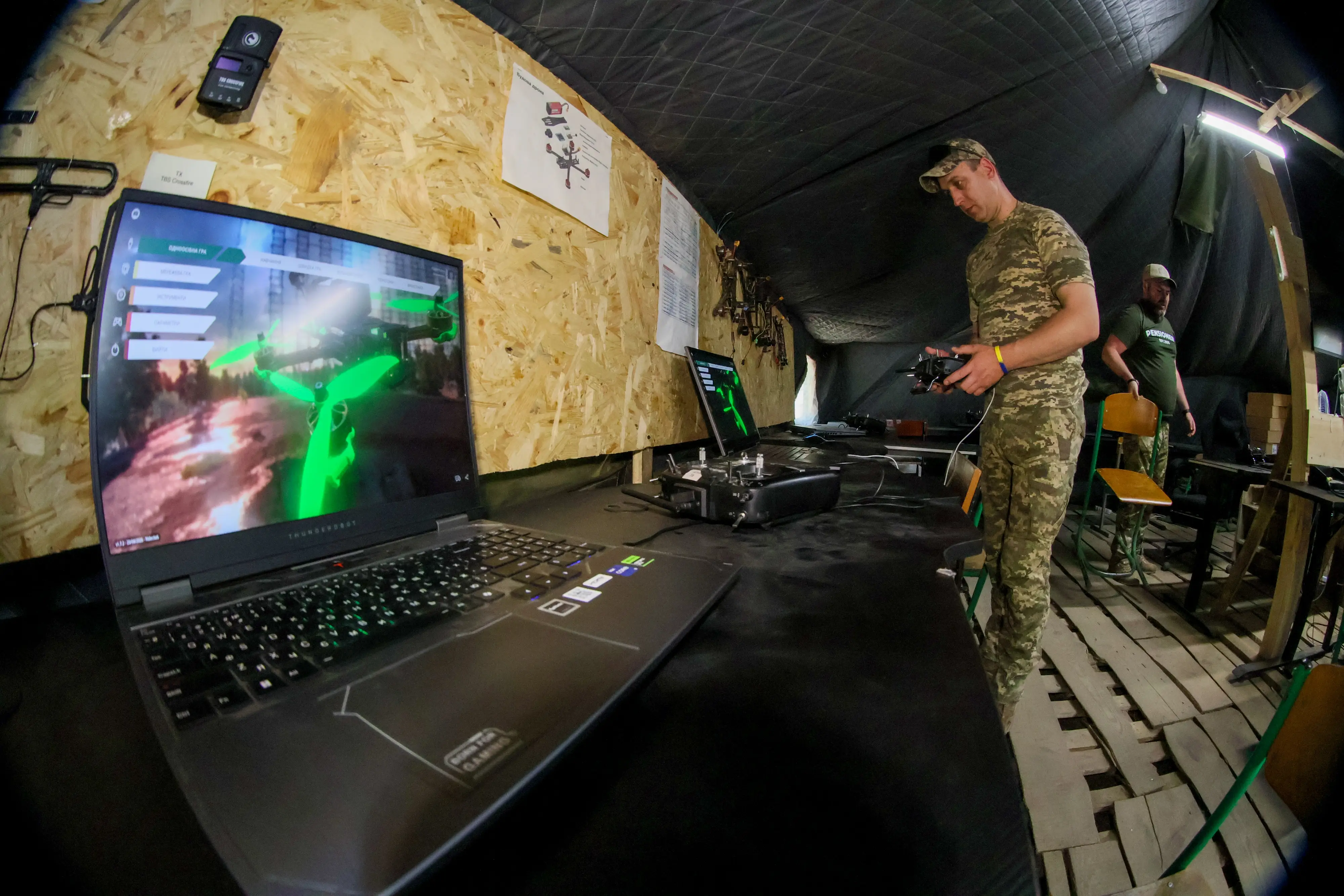 Person in camouflage uniform operates a controller beside laptops and drone equipment inside a tented workstation.