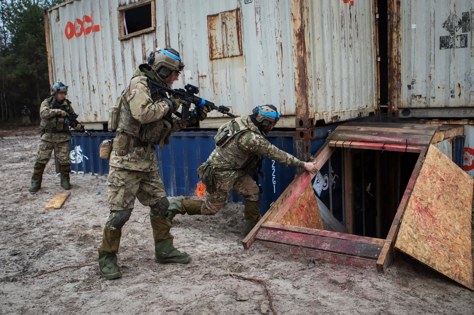 Camouflage-clad tactical personnel with rifles move around stacked shipping containers during an outdoor training exercise.