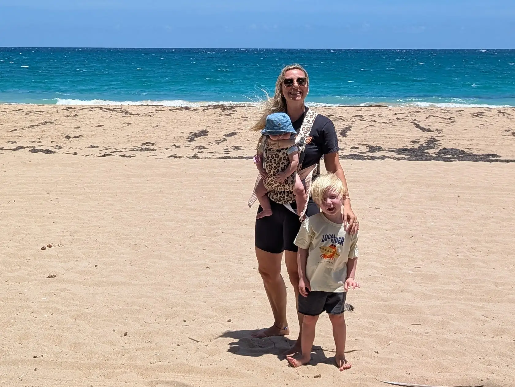 Woman with baby in carrier smiling with toddler on beach
