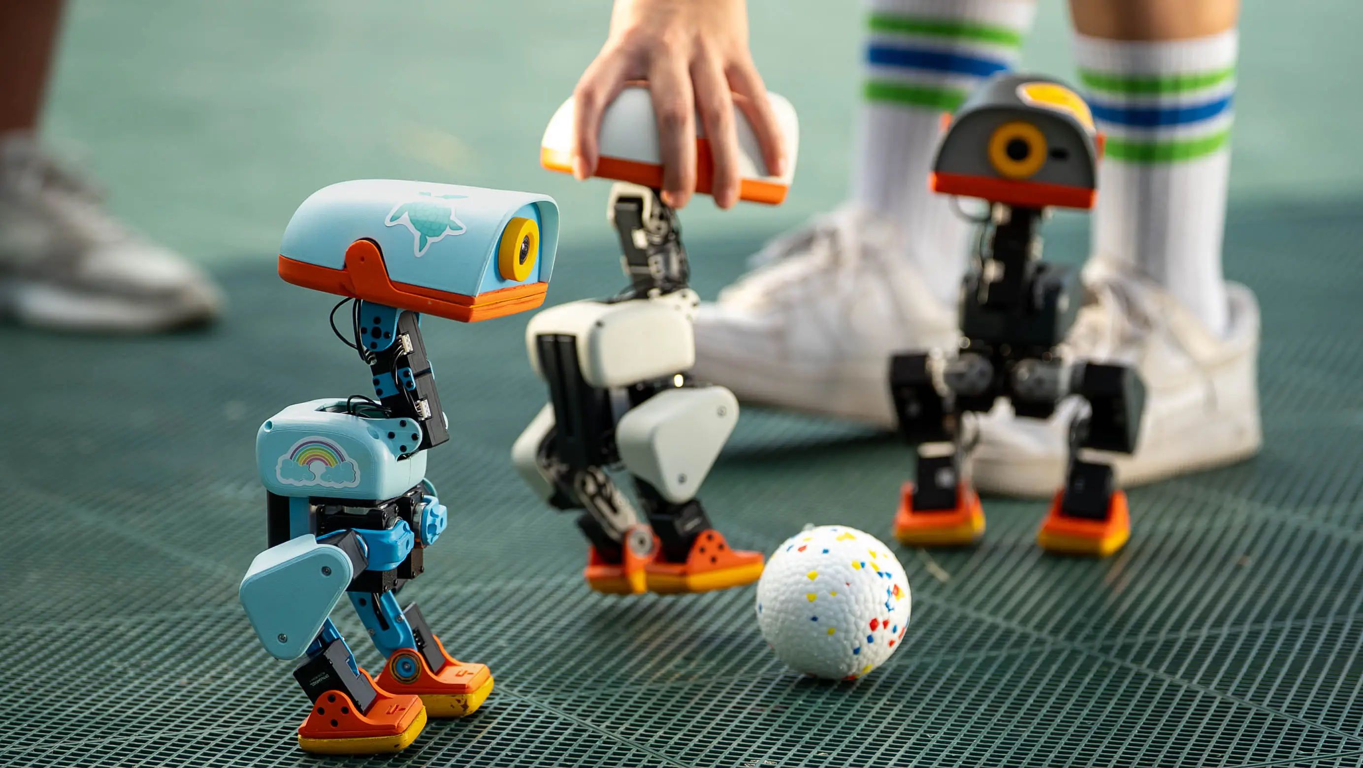 Three Hugging Face and Pollen Robotics Microduck robots stand on a green platform with two humans in the background. Two of the Microducks are looking at a white ball.