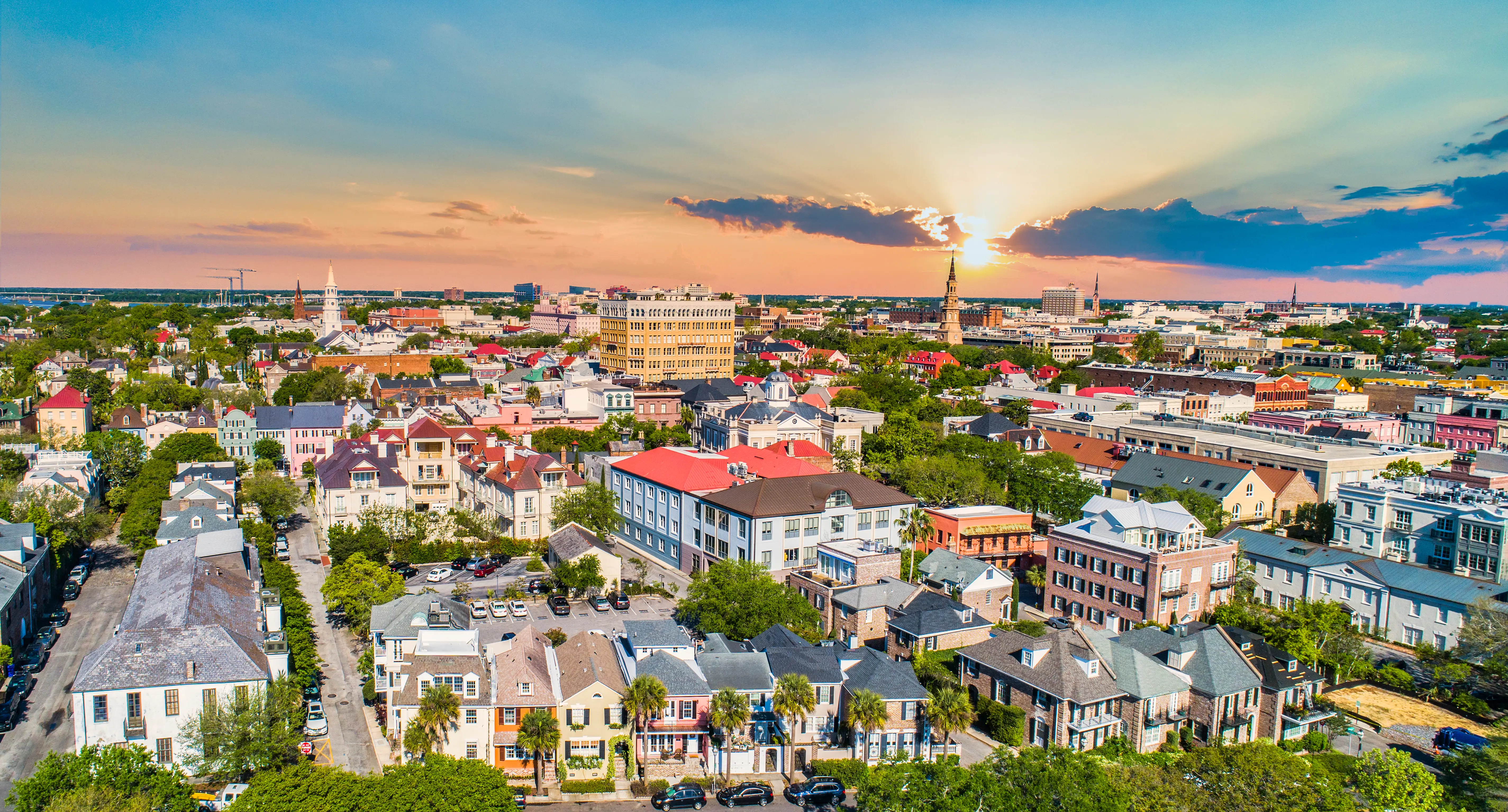 Aerial view of historic Charleston rooftops and church steeples at sunset under a colorful sky.