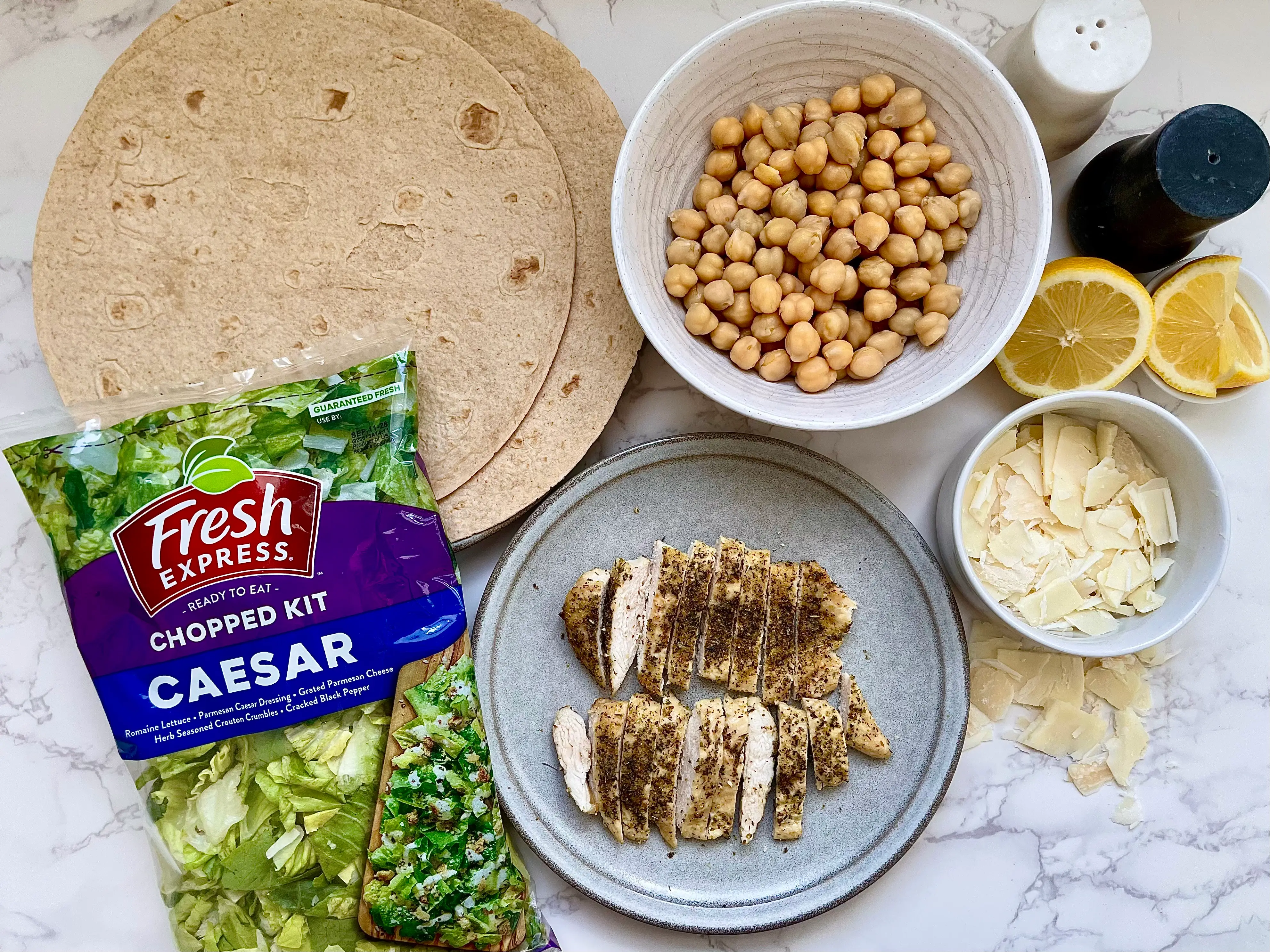 A Caesar salad kit, tortillas, chickpeas, grilled chicken, Parmesan cheese, lemons, salt, and pepper on a countertop.