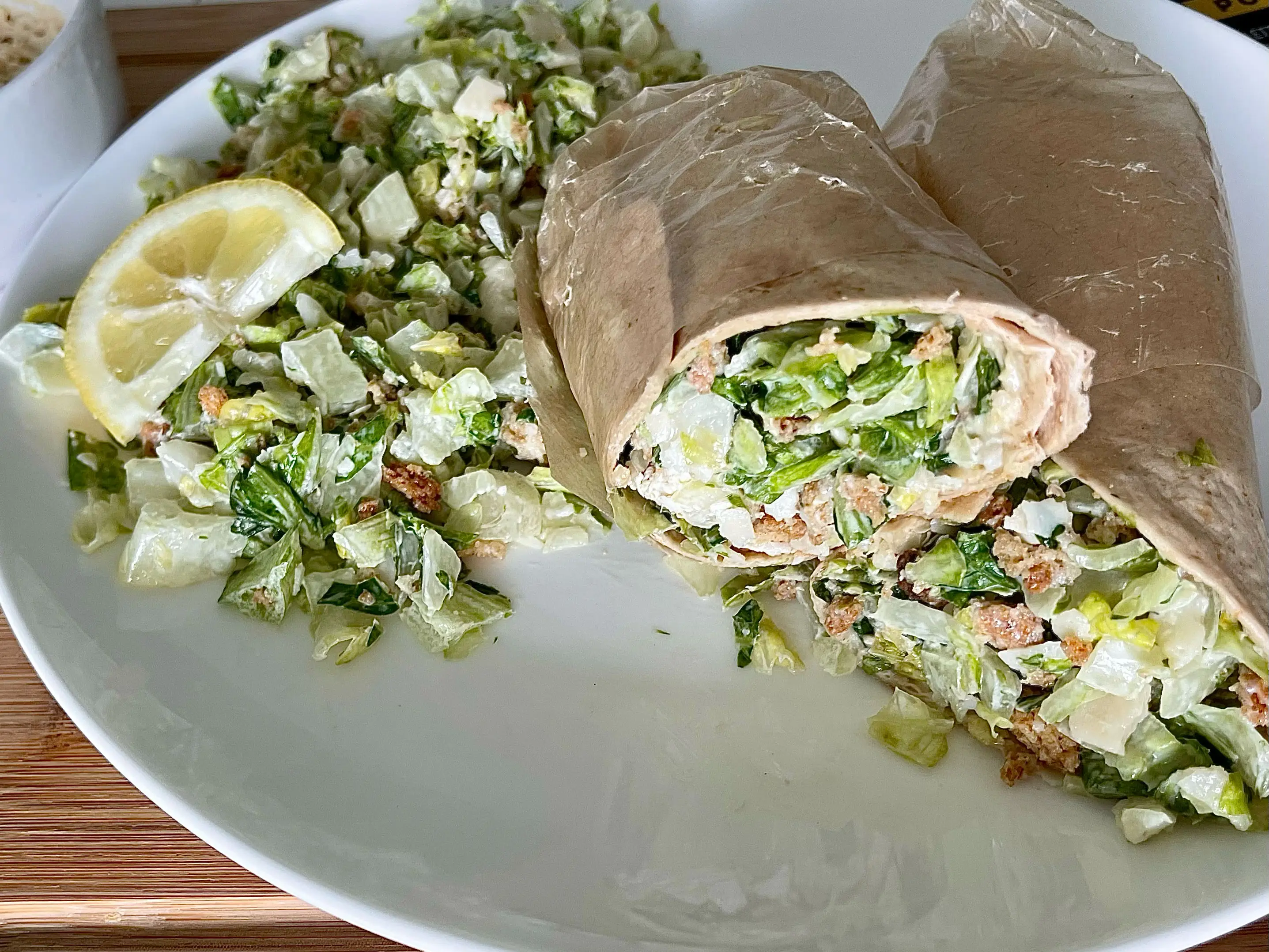 A grilled chicken Caesar wrap on a white plate with extra salad and a lemon on the side.