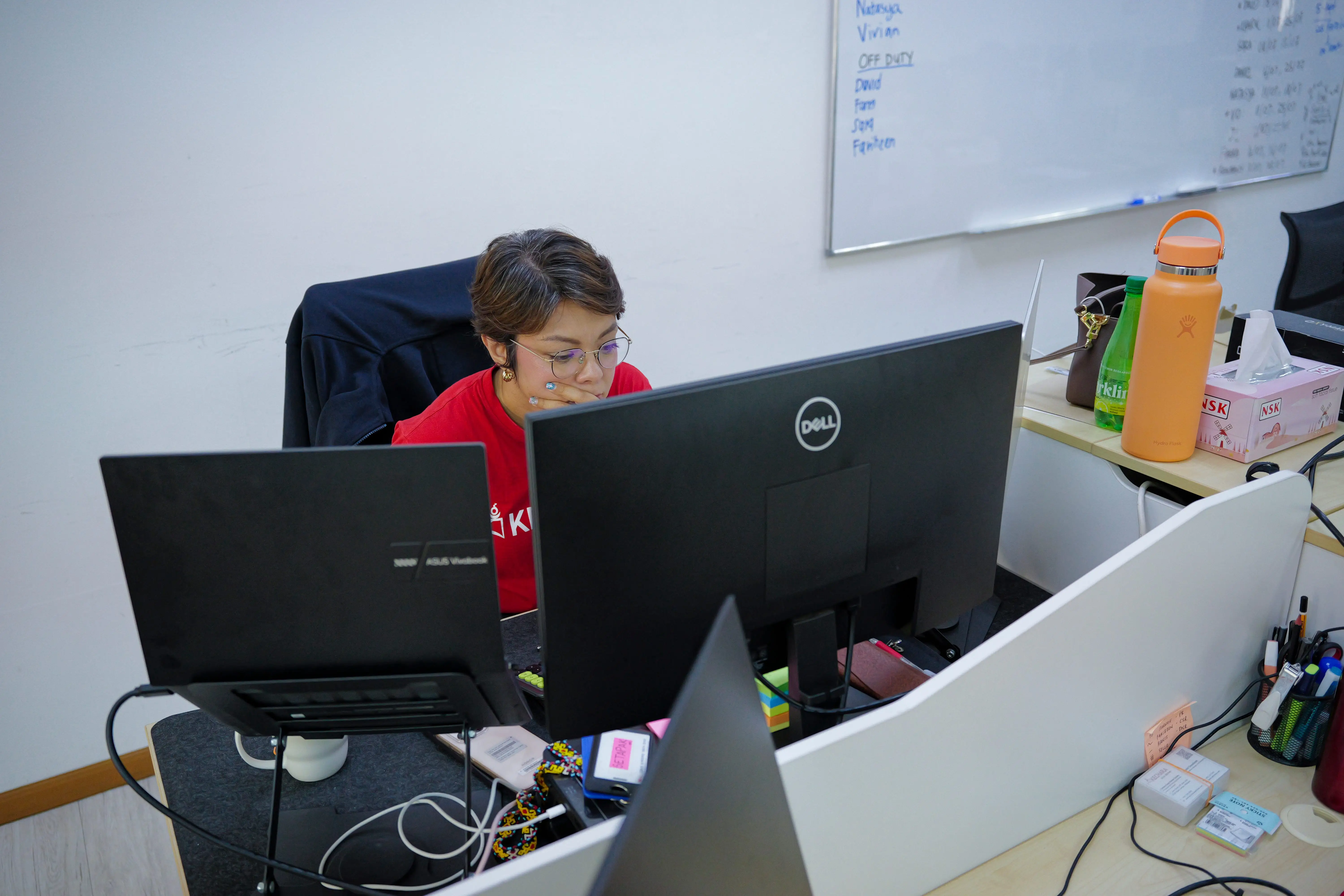 Office worker at a desk uses multiple screens in front of a whiteboard and office supplies.