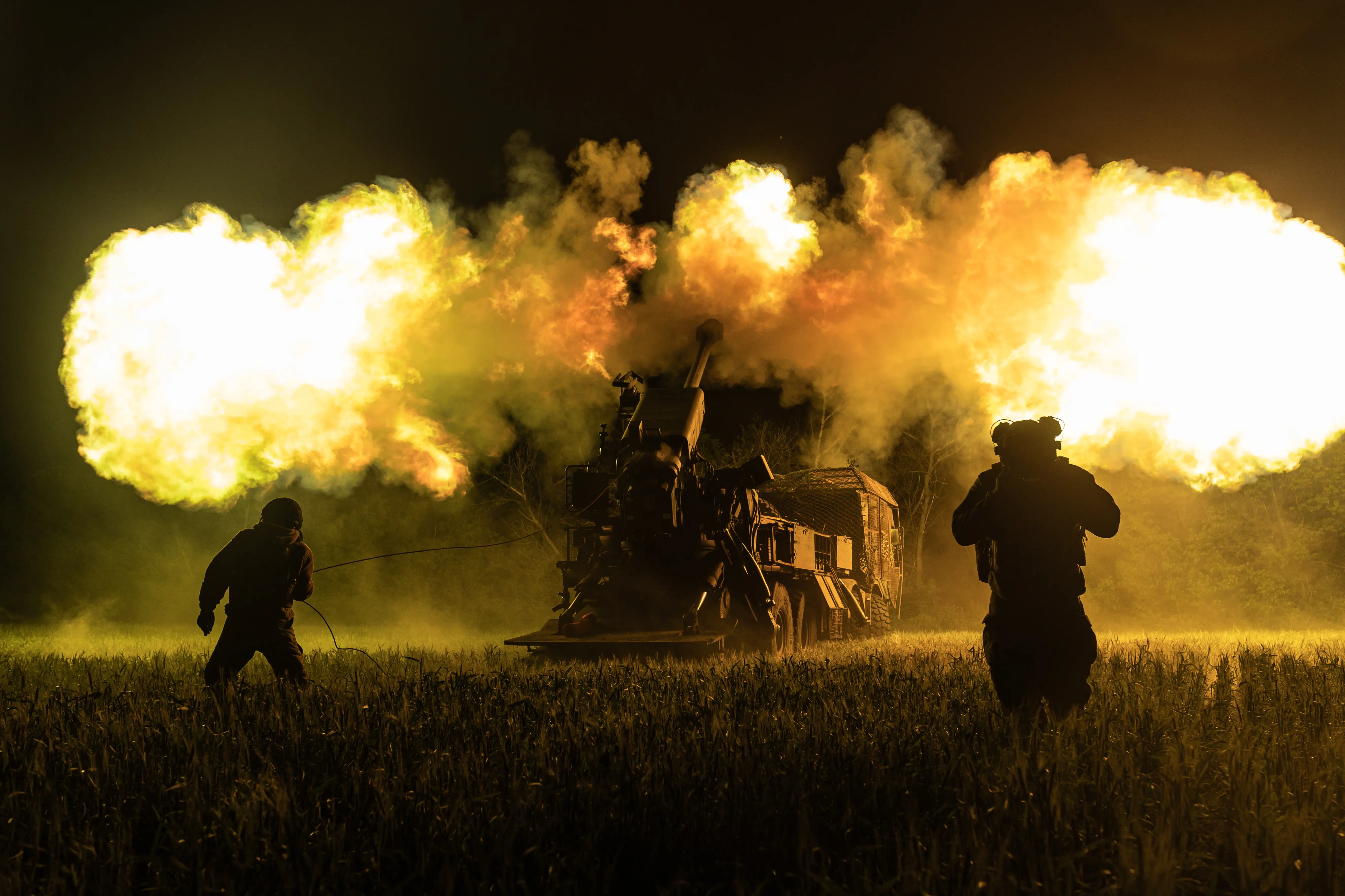 Silhouetted soldiers stand near artillery firing large blasts across a dark field at night.