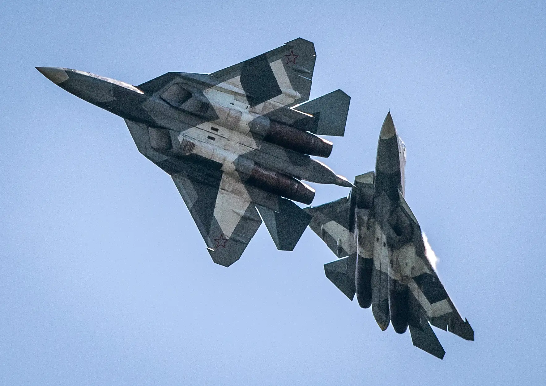 Two Sukhoi Su-57 fighter jets with camouflage paint and red star markings fly close together against a blue sky.