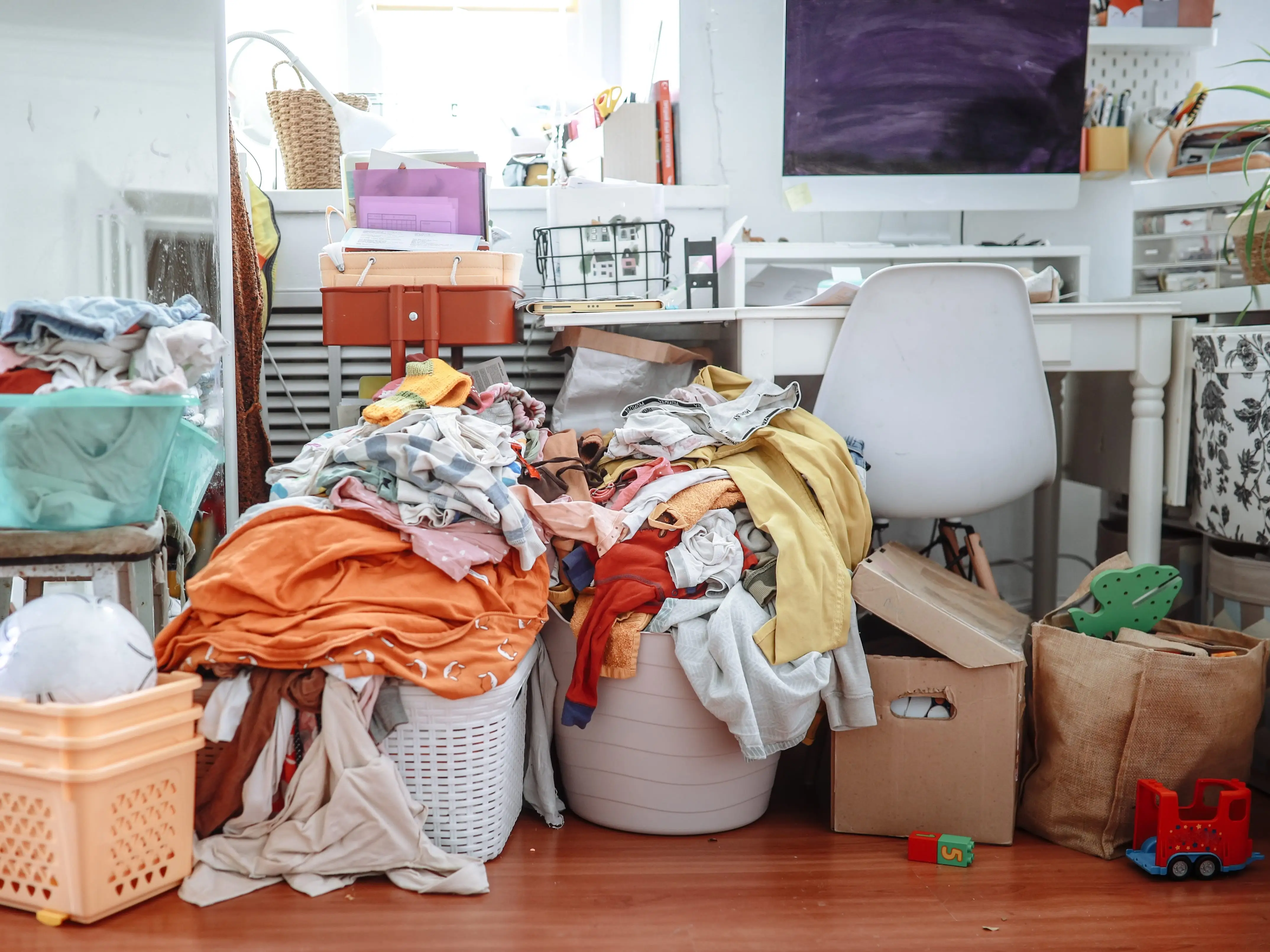 Cluttered room with piles of laundry, storage bins, toys, and a desk with a white chair.