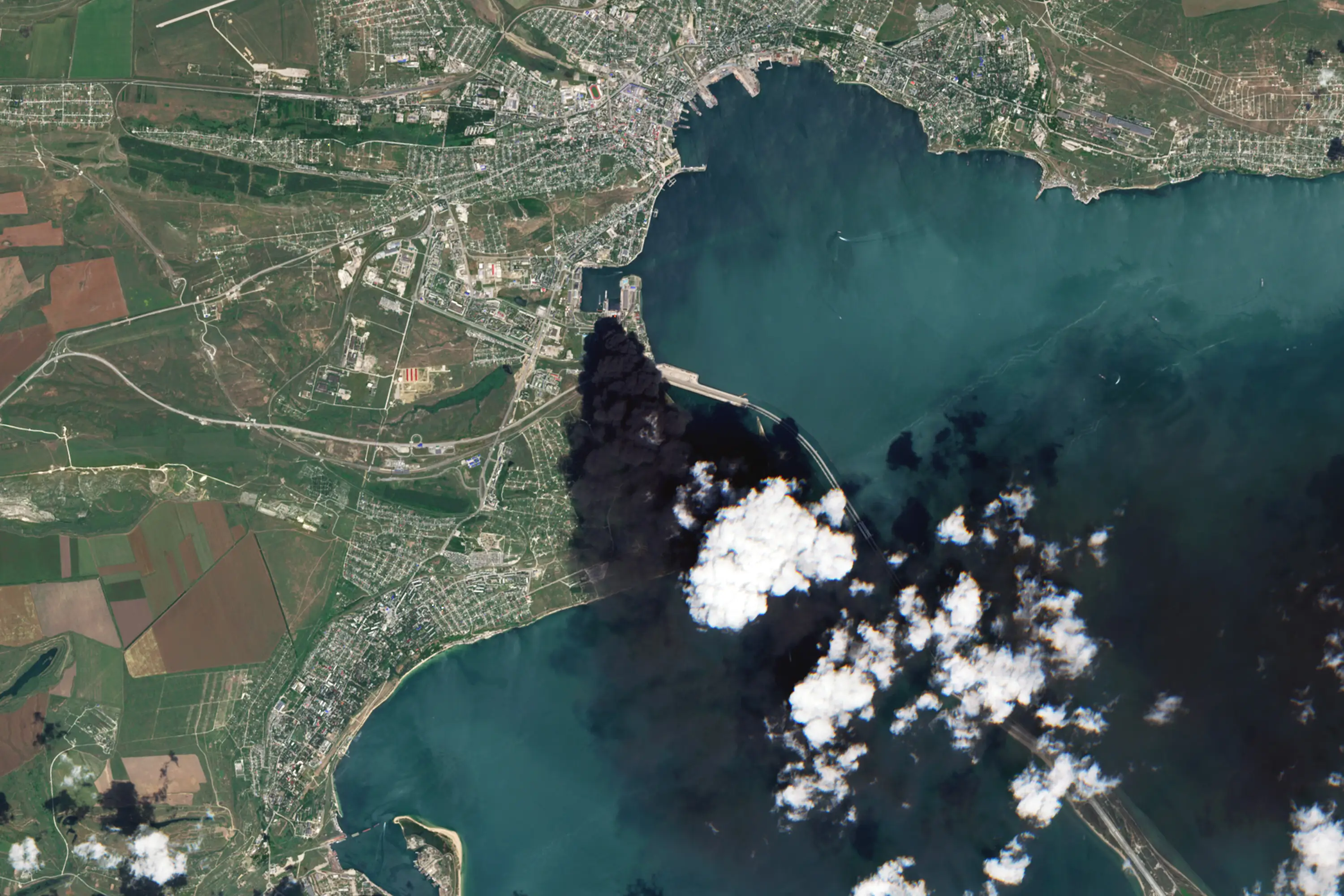 Satellite view shows a coastal harbor city with dense black smoke rising from a waterfront area.
