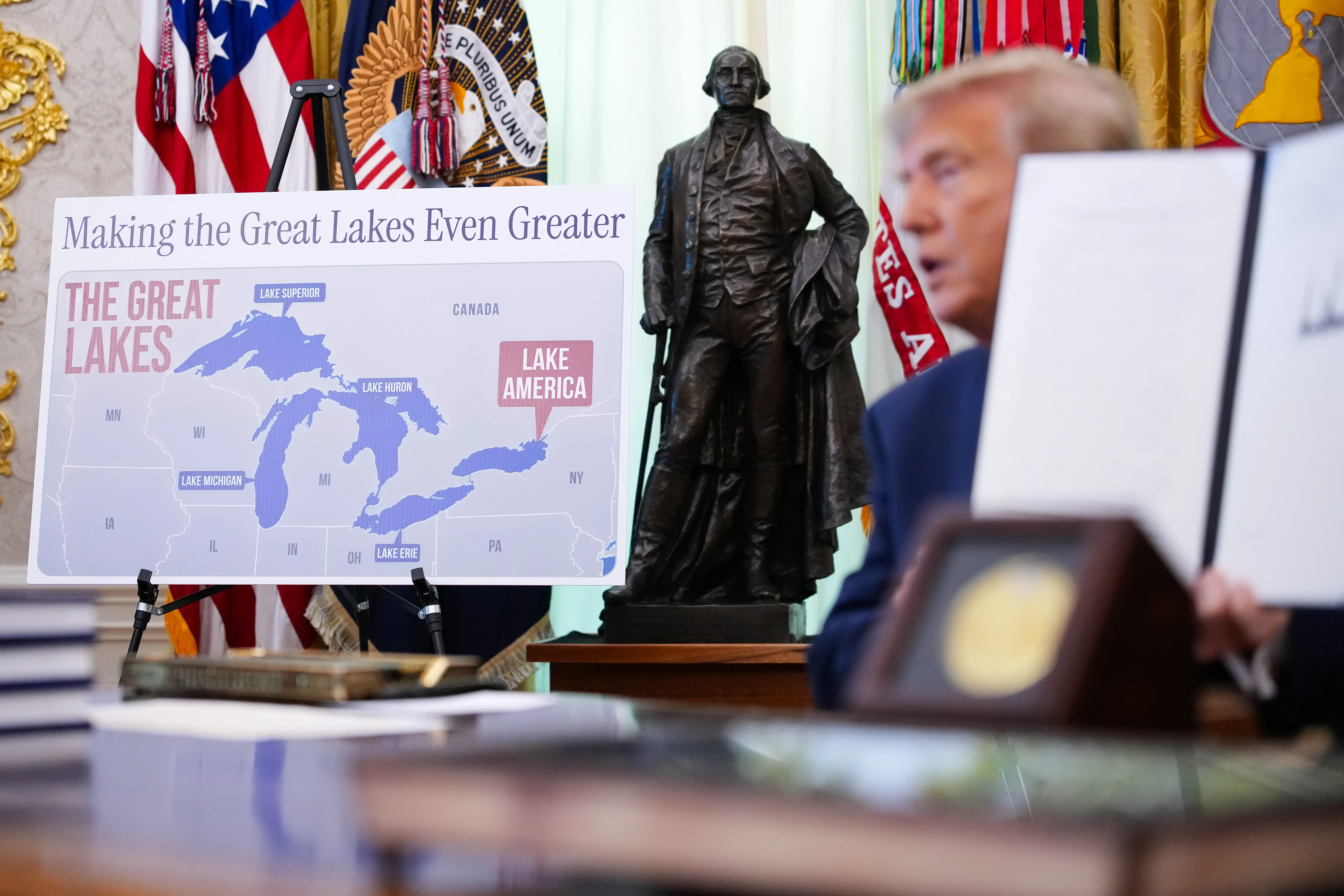 President Donald Trump sits at the Resolute Desk with a map of the Great Lakes, including the eastern-most lake labeled
