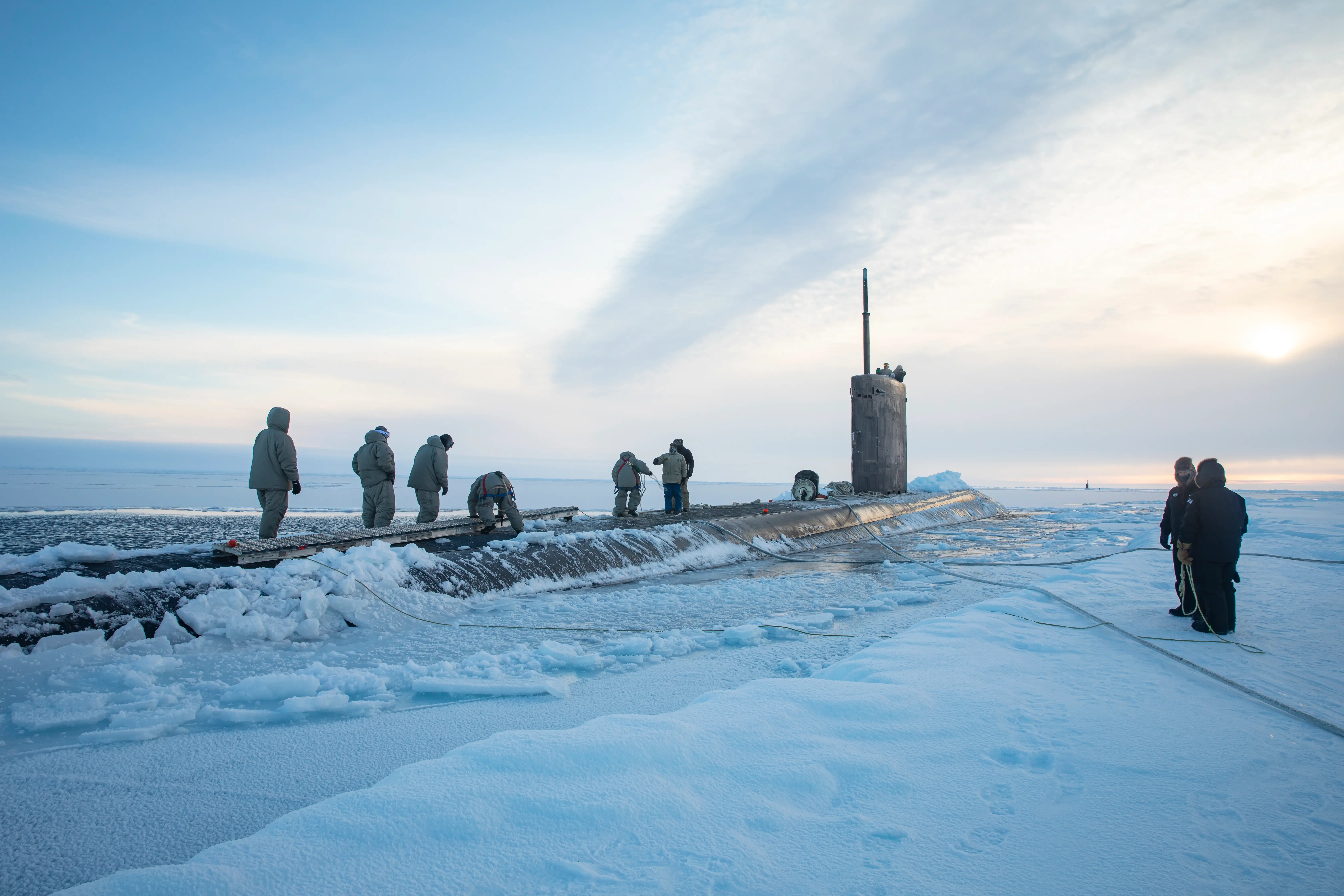 The USS Pasadena submarine sails in icy Arctic waters. A group of men stand on top of it and on ice nearby.