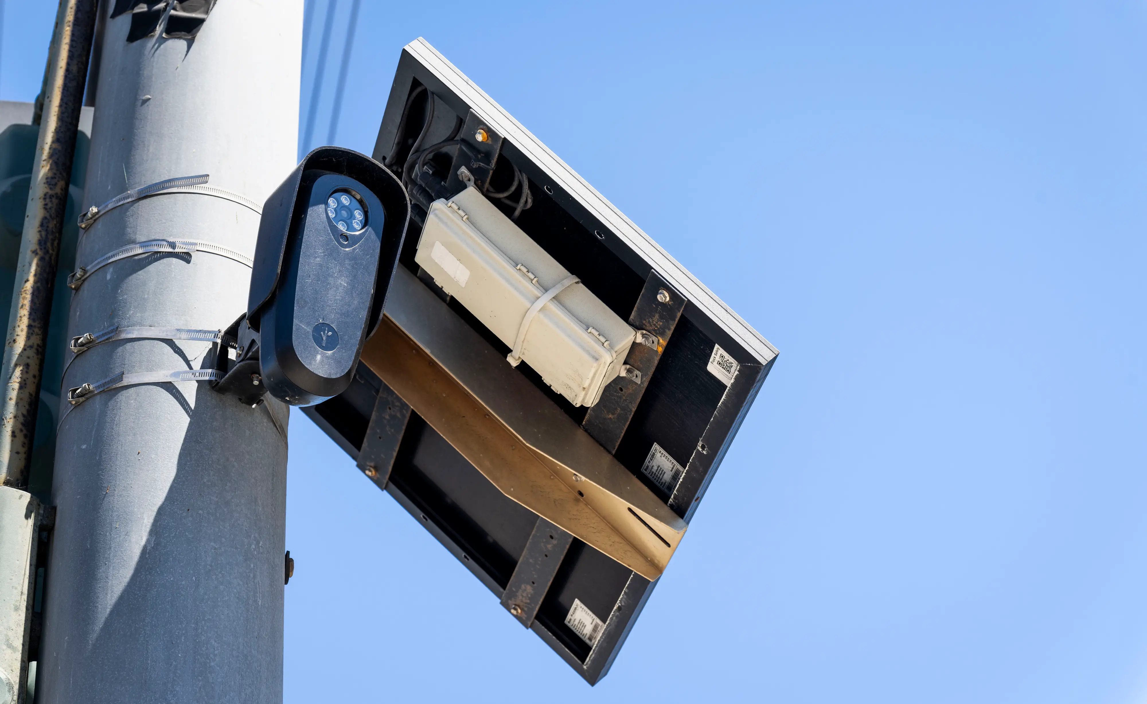 Traffic monitoring camera mounted on a metal pole beside an open equipment box against a clear blue sky.