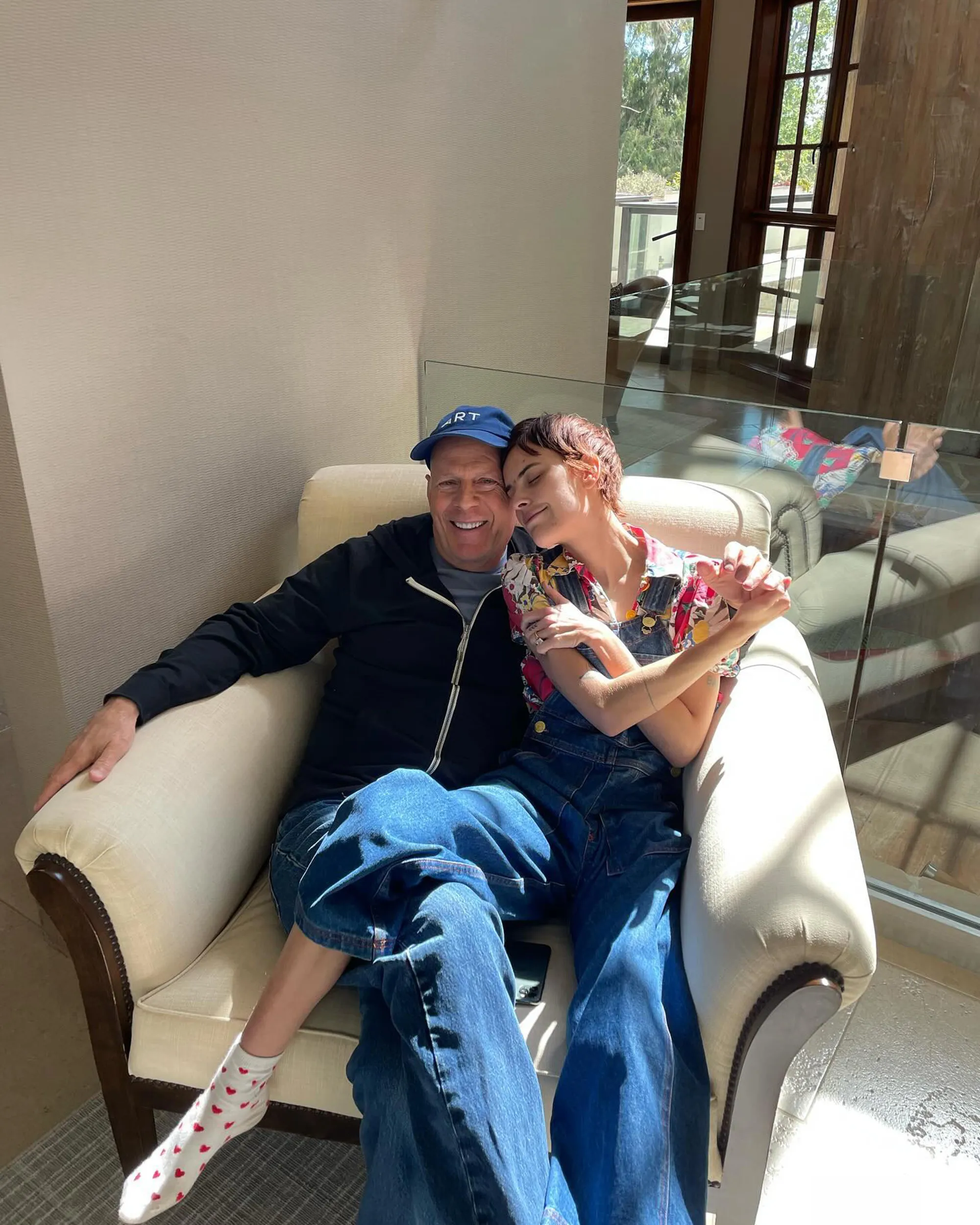 Bruce Willis and daughter Tallulah Willis sitting together in an armchair, smiling, with Tallulah resting her head on her father's shoulder.