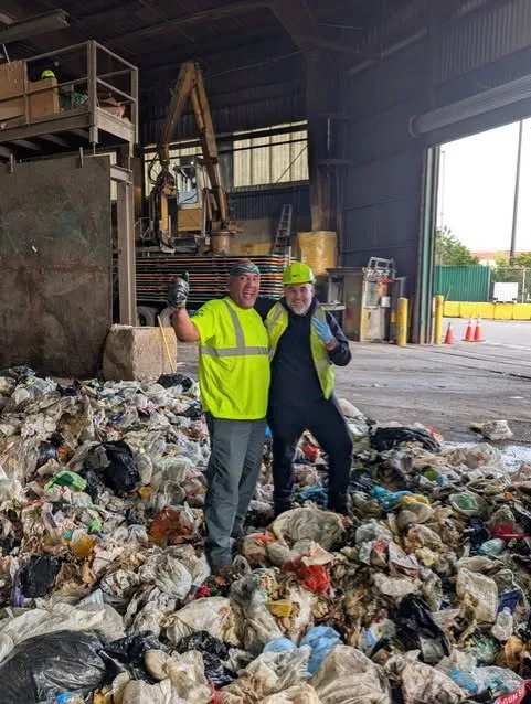 Two workers in high-visibility vests standing on a large pile of trash in a processing facility.
