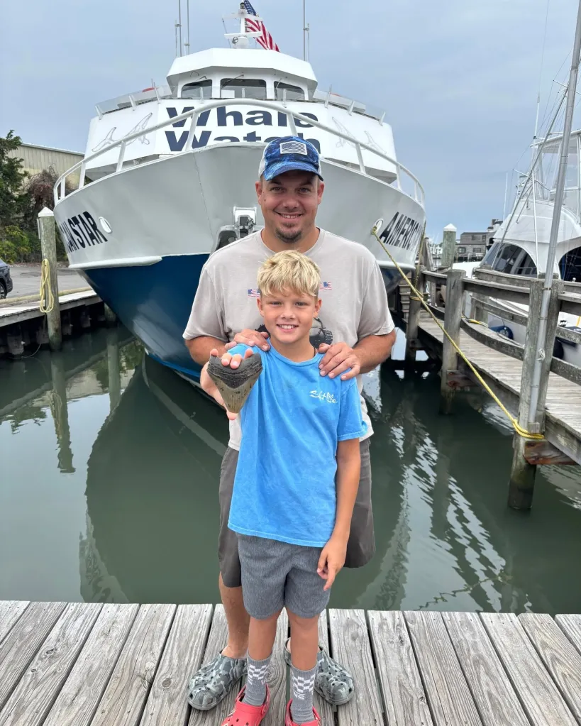 A man and a boy stand on a dock, holding up a large megalodon tooth.