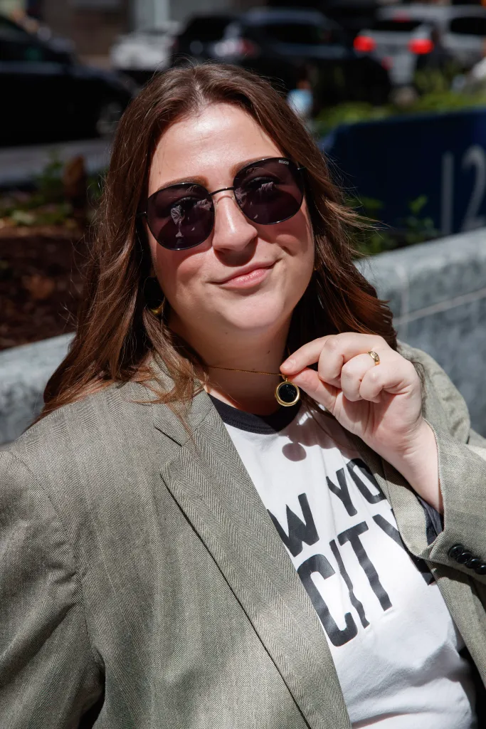 McKenzie wearing sunglasses and a blazer, holding up a UV necklace.