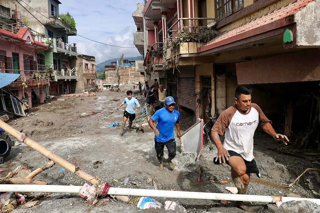 People navigate a street covered in mud and debris after flash floods, with damaged buildings in the background.