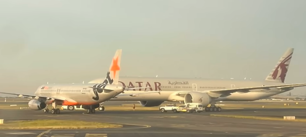 A Jetstar plane is seen in the foreground with its tail fin and a portion of its body, while a large Qatar Airways plane is visible in the background, partially obscured by the smaller aircraft.