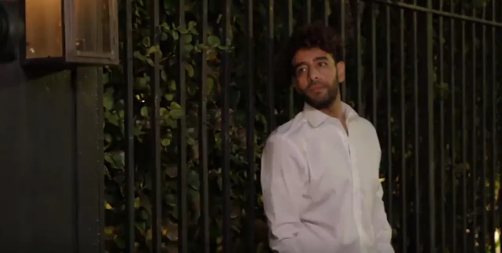 Mido, a man with curly brown hair and a beard, wearing a white button-down shirt, stands outside against a dark fence covered in green leaves.
