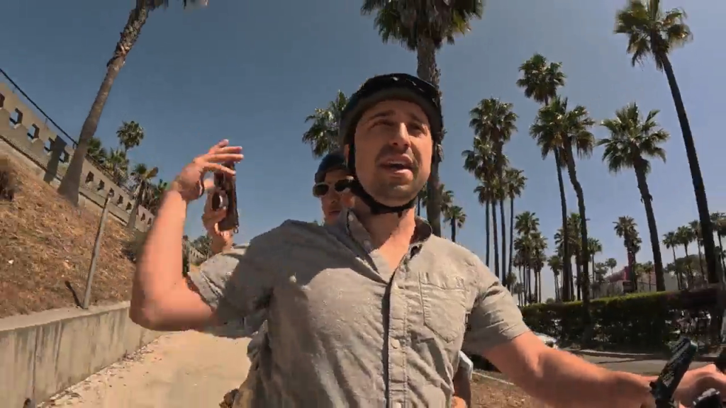 A man in a helmet and shirt holding a phone while riding a bike with another person behind him on a sunny street lined with palm trees.