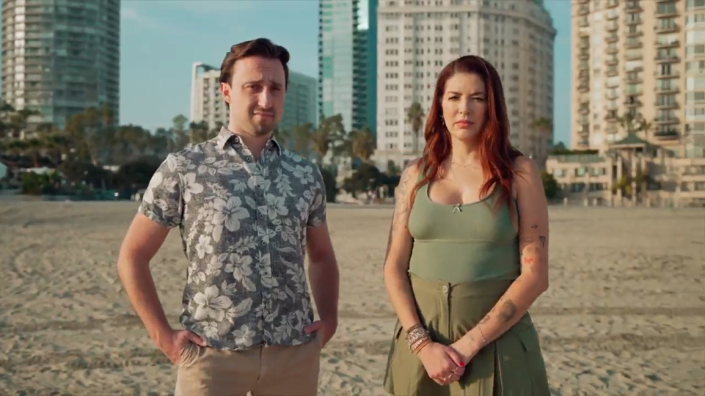 A man and a woman stand on a sandy beach with city buildings in the background.