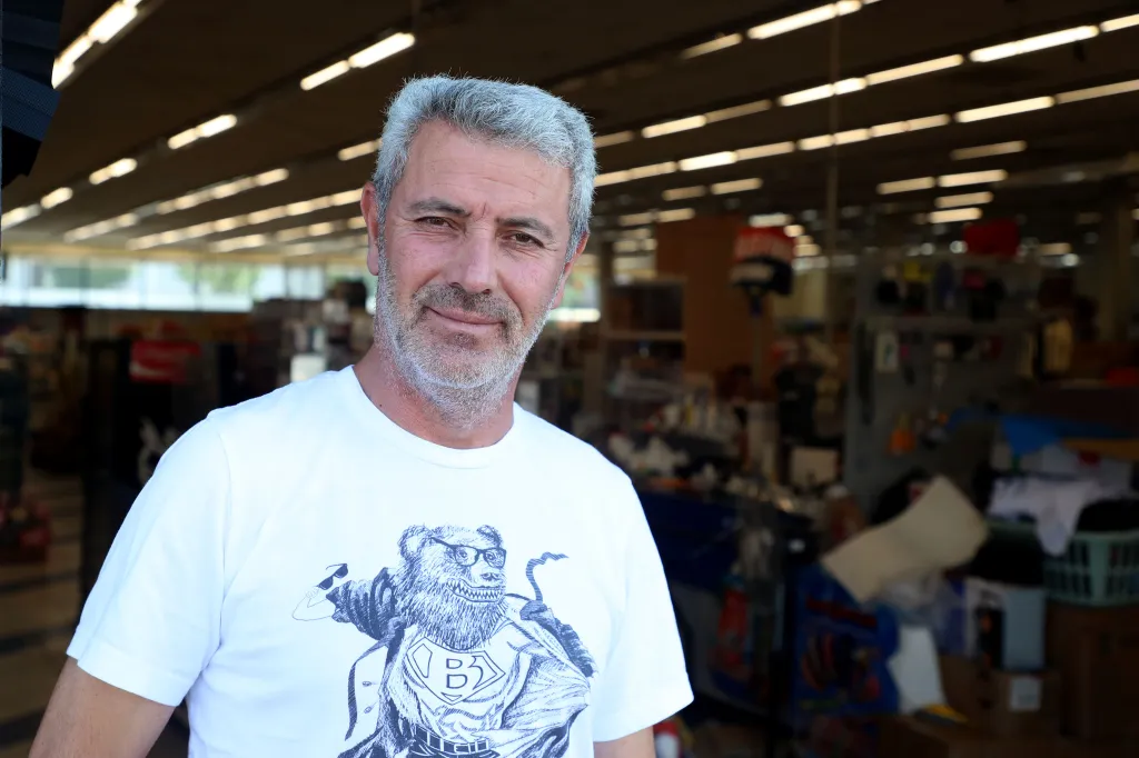 A man, identified as a 99c store worker, smiles faintly in front of the store with visible merchandise in the background.