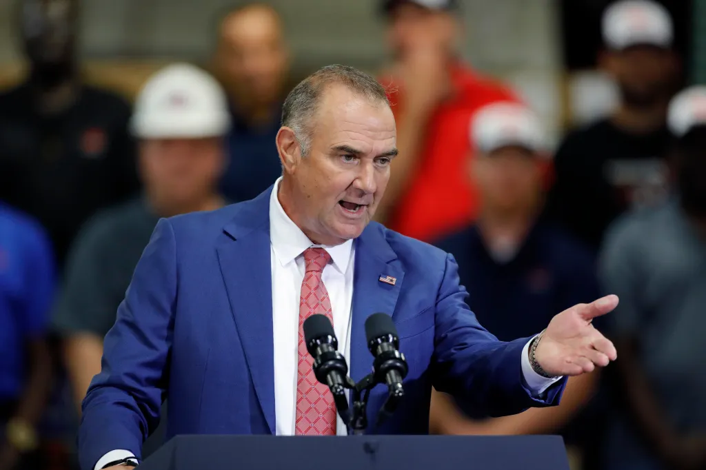 Man in a blue suit and red tie speaking at a podium with microphones, gesturing with his right hand.