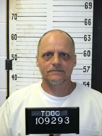 Mugshot of a balding white man with a goatee in a white shirt, with a board around his neck reading 