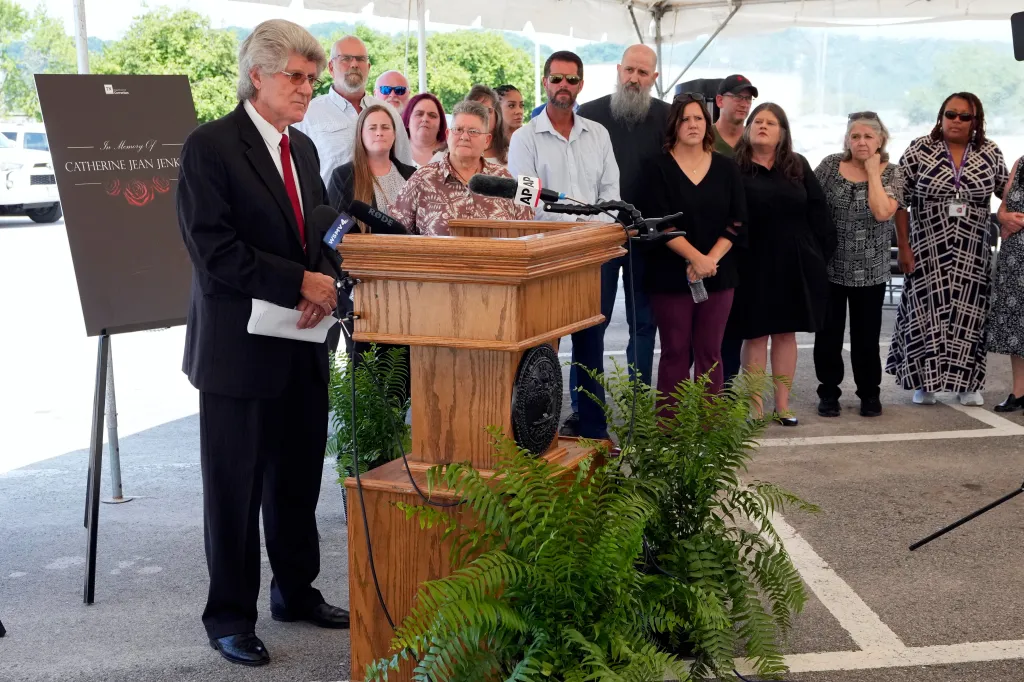 Dennis Jenkins at a podium with family members, speaking under a tent.