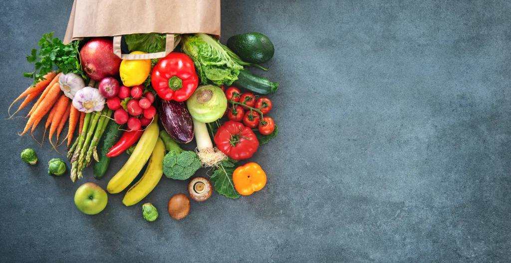 A paper shopping bag overflowing with a variety of fresh vegetables and fruits, including carrots, bell peppers, tomatoes, bananas, and a pomegranate, against a dark gray background.