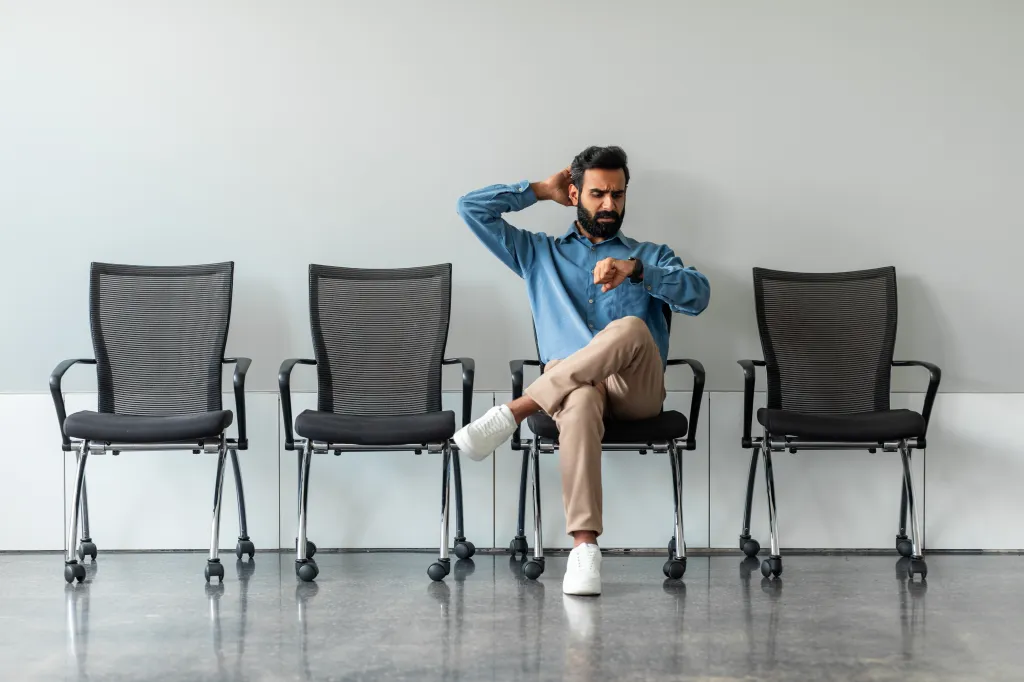 A worried Indian man sitting alone in a waiting room, checking his watch impatiently.