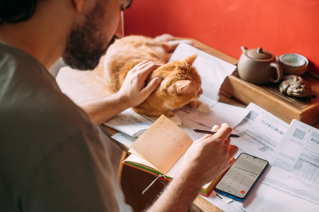 A man calculating utility bills with a smartphone, notebook, and pen, while petting an orange cat.