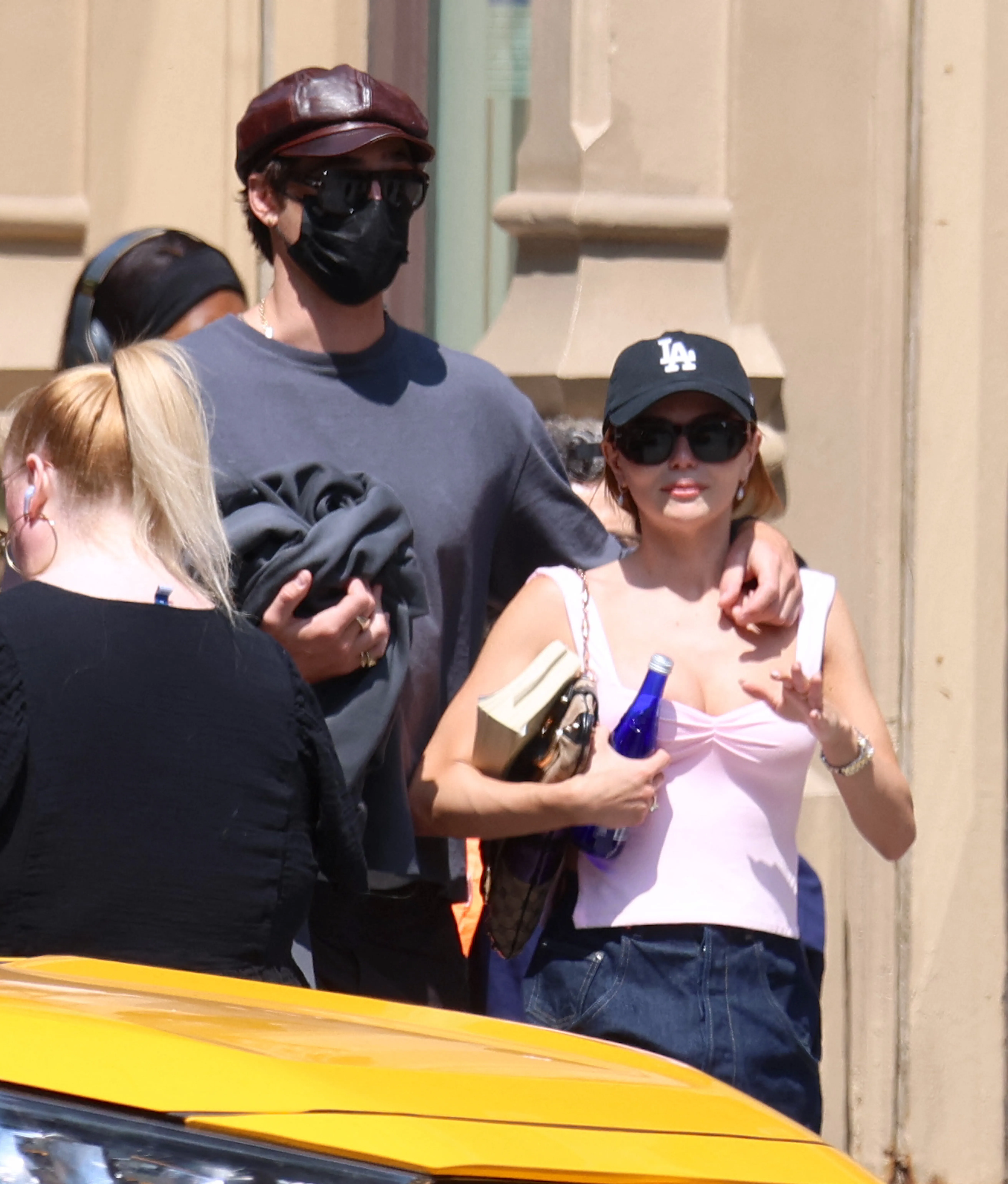 A man in a brown leather cap and black face mask walks with his arm around a woman in a black LA baseball cap and pink top.