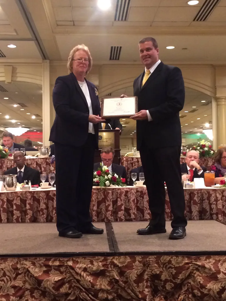 A woman in a navy suit presenting a framed certificate to a man in a black suit and yellow tie on a stage.