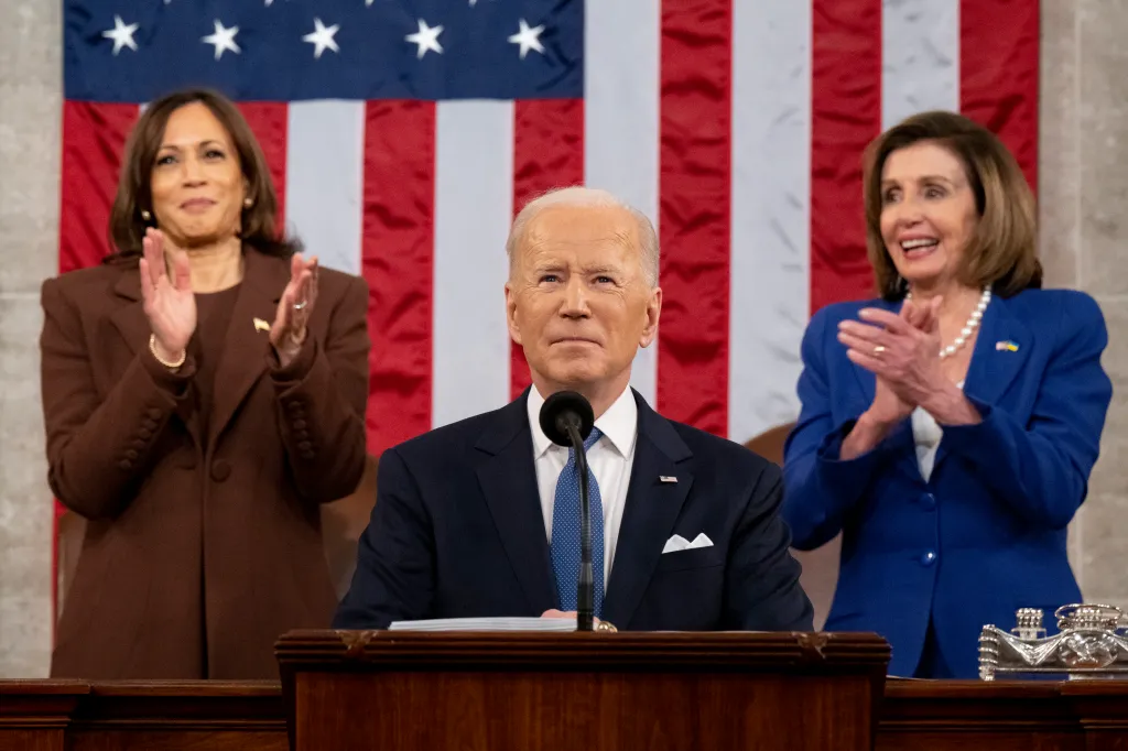 President Joe Biden stands at a podium while Vice President Kamala Harris and Speaker of the House Nancy Pelosi clap behind him with an American flag background.
