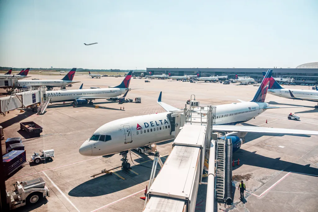 Several Delta airplanes parked at airport gates on a sunny day, with one plane taking off in the background.