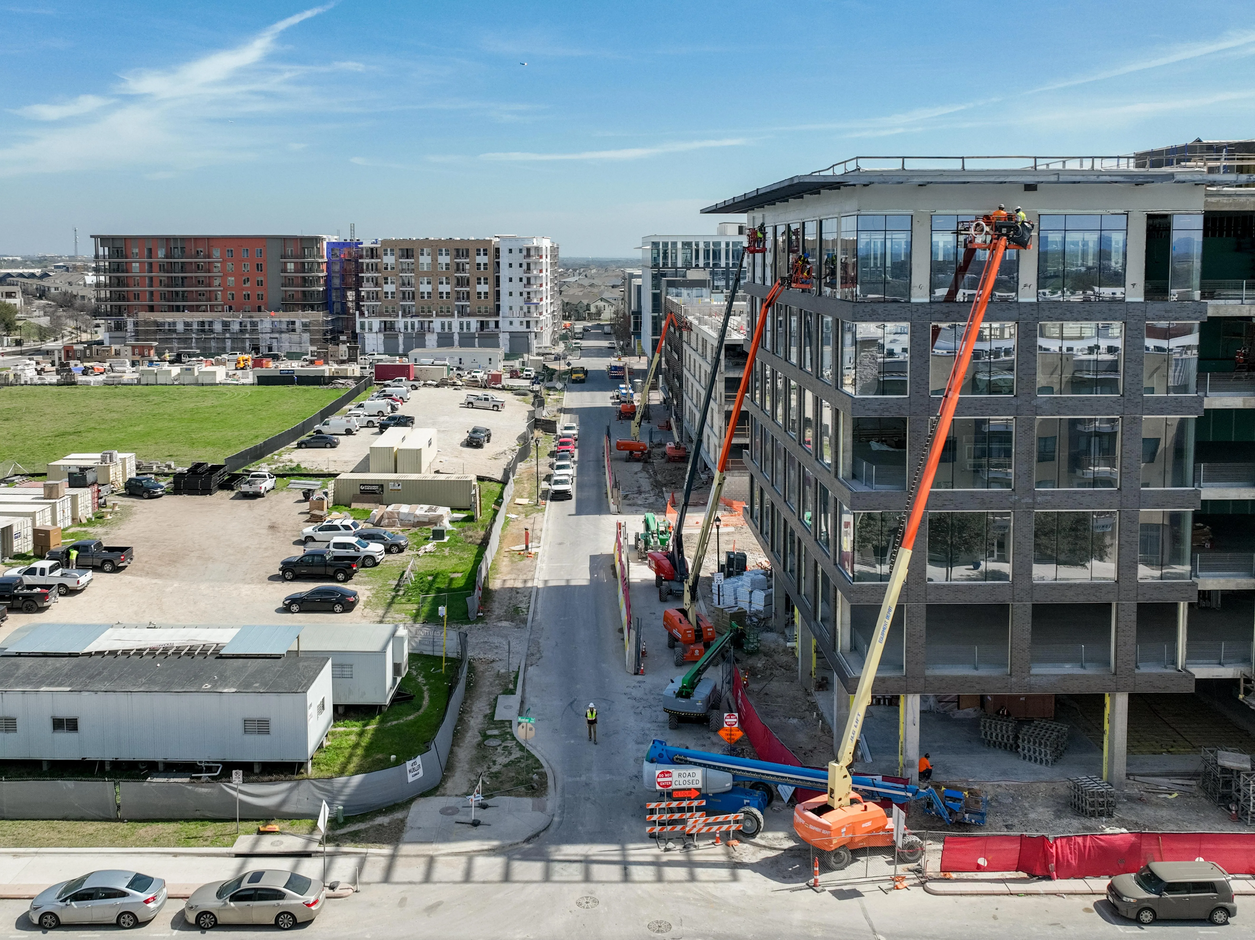 Construction workers on lifts install windows on a new mid-rise apartment building, with other recently built apartment complexes visible nearby.