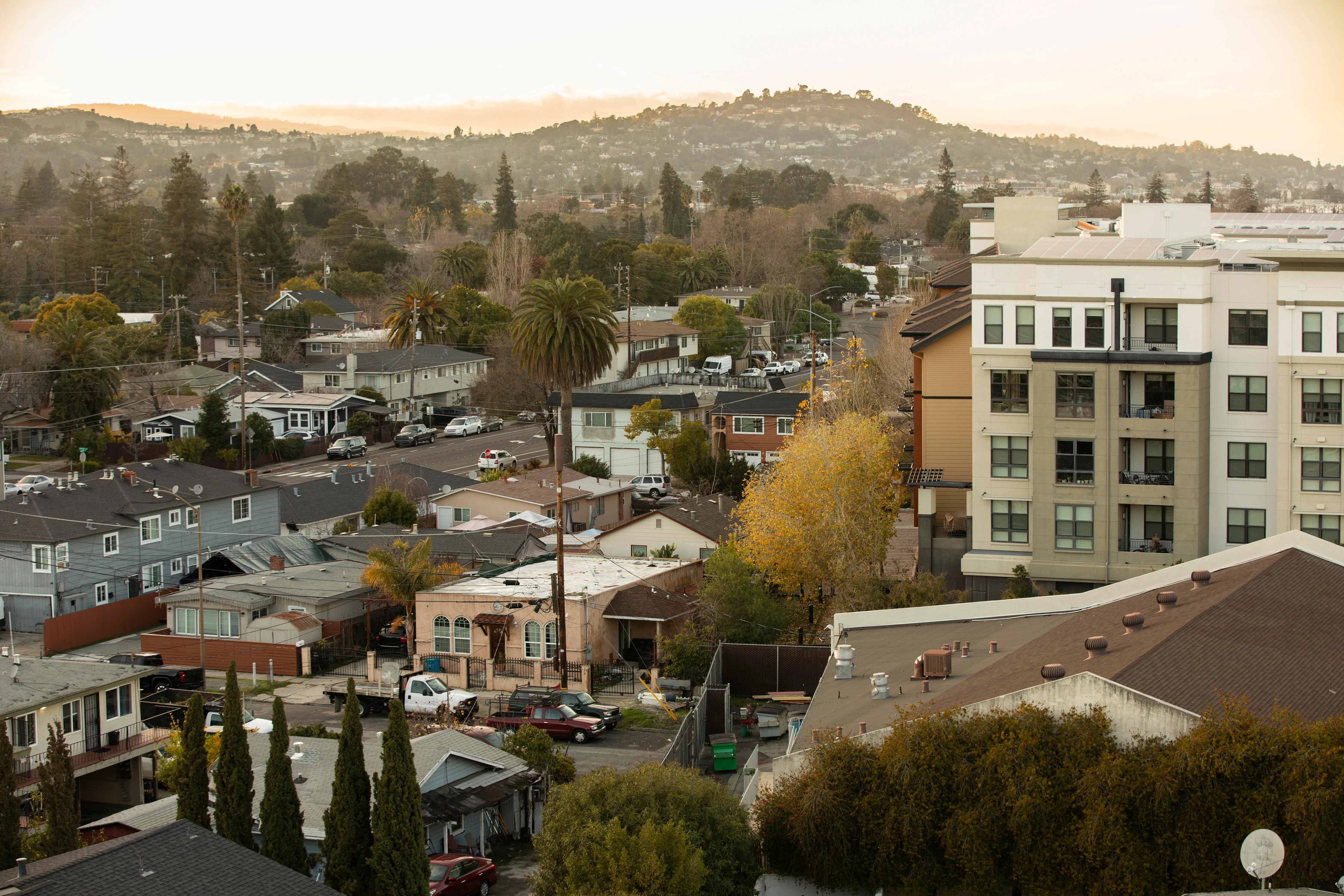 Single-family homes and a mid-rise apartment building fill a residential neighborhood in Redwood City, California, with hills in the distance.