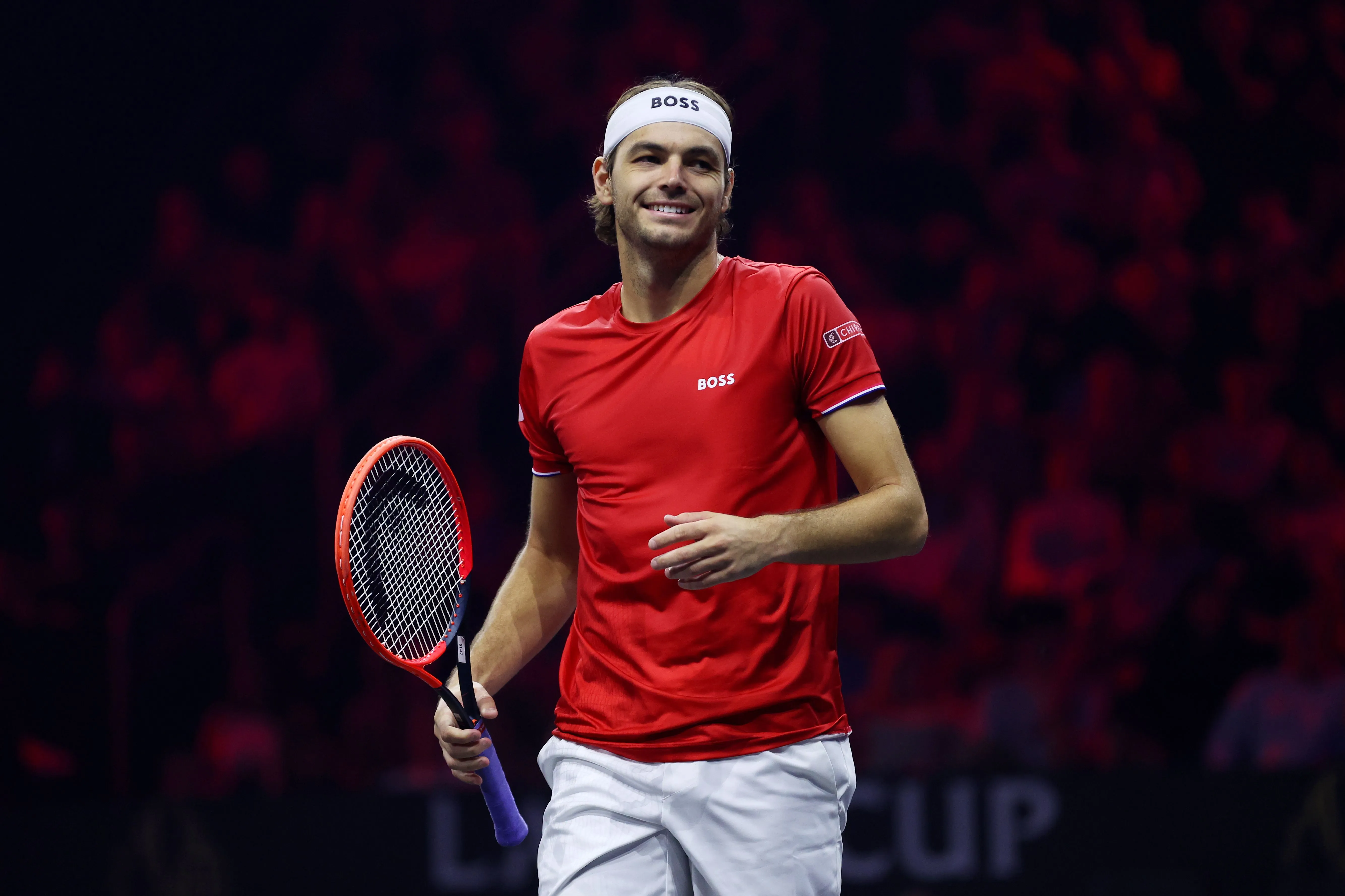Taylor Fritz smiling in a red shirt and white headband, holding a tennis racket.