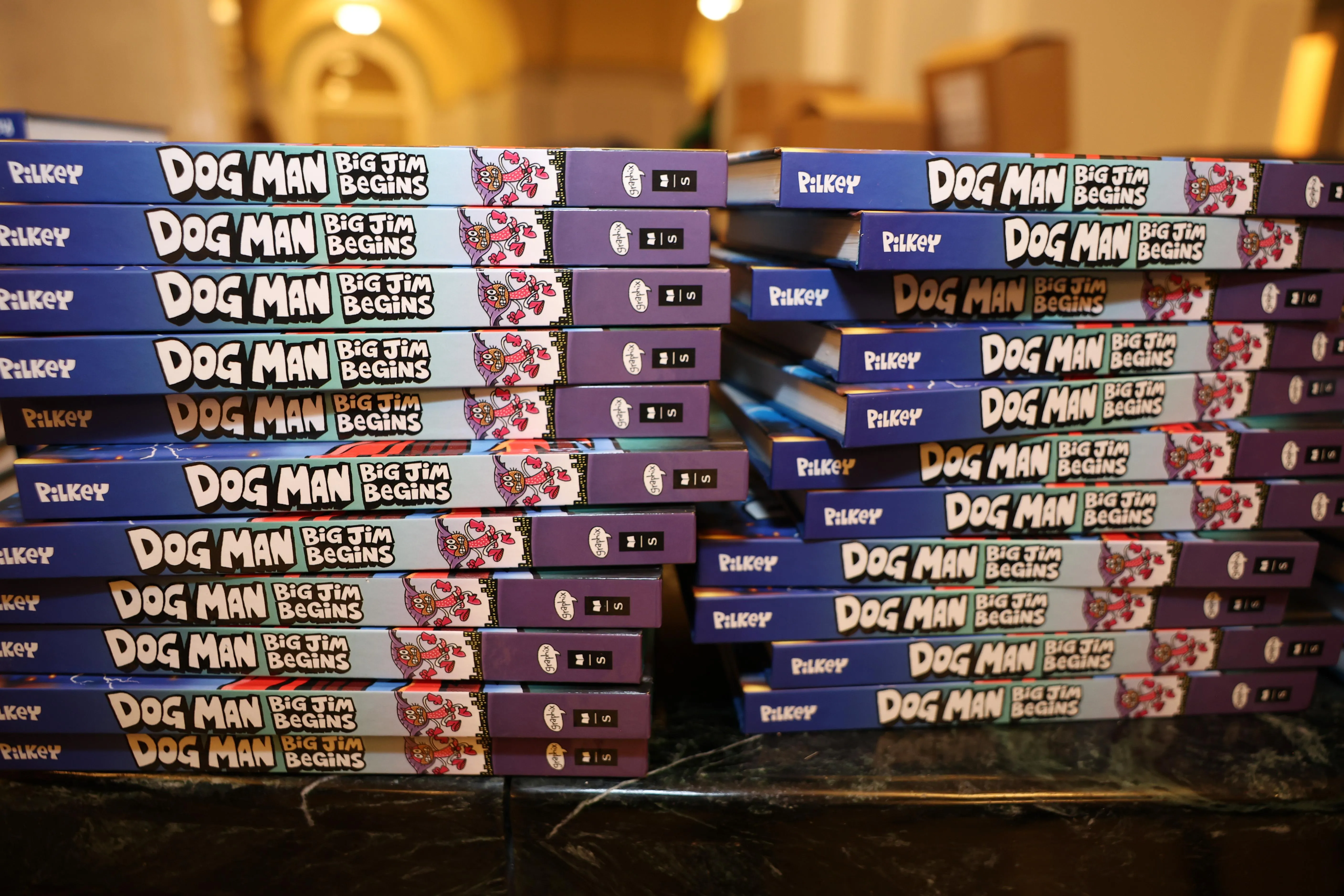 Two stacks of books lie on a table, their spines toward the camera. They’ll all copies of the same book, entitled DOG MAN: BIG JIM BEGINS, in cartoonish bubble letters. Two stacks of books lie on a table, their spines toward the camera. They’ll all copies of the same book, entitled DOG MAN: BIG JIM BEGINS, in cartoonish bubble letters.