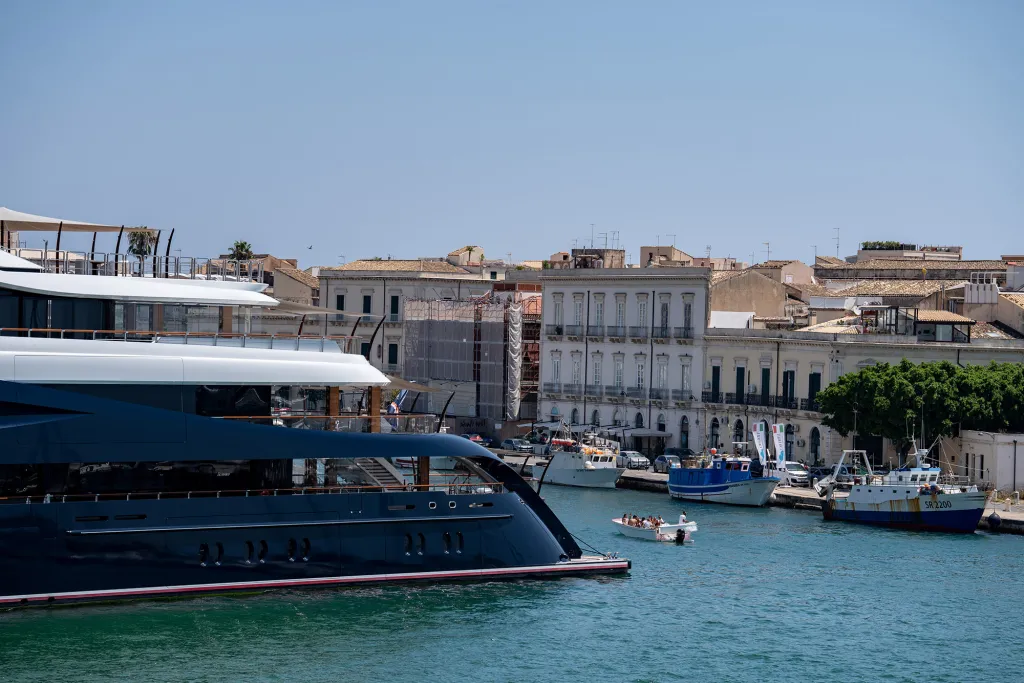 A dark blue yacht docked in a harbor with a town in the background.