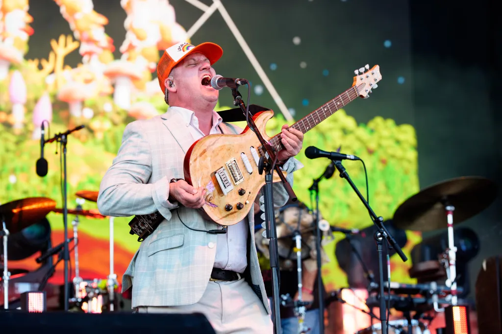 A man in an orange hat, white shirt, and light gray blazer sings into a microphone while playing an electric guitar.