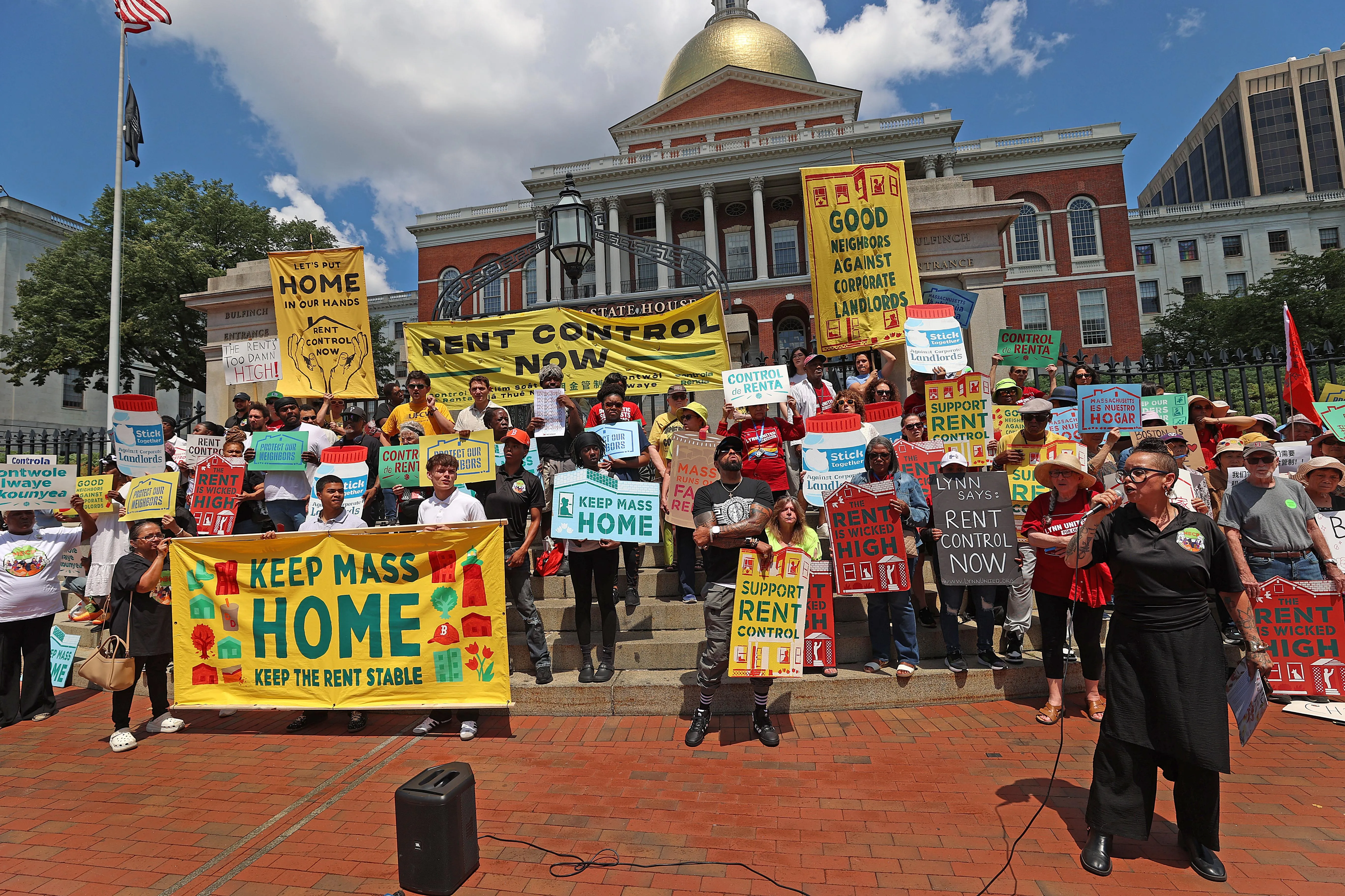 A crowd of rent-control supporters gathers on the steps of the Massachusetts State House, holding colorful signs that read “Rent Control Now,” “Keep Mass Home,” and “Support Rent Control,” as a speaker addresses the rally.