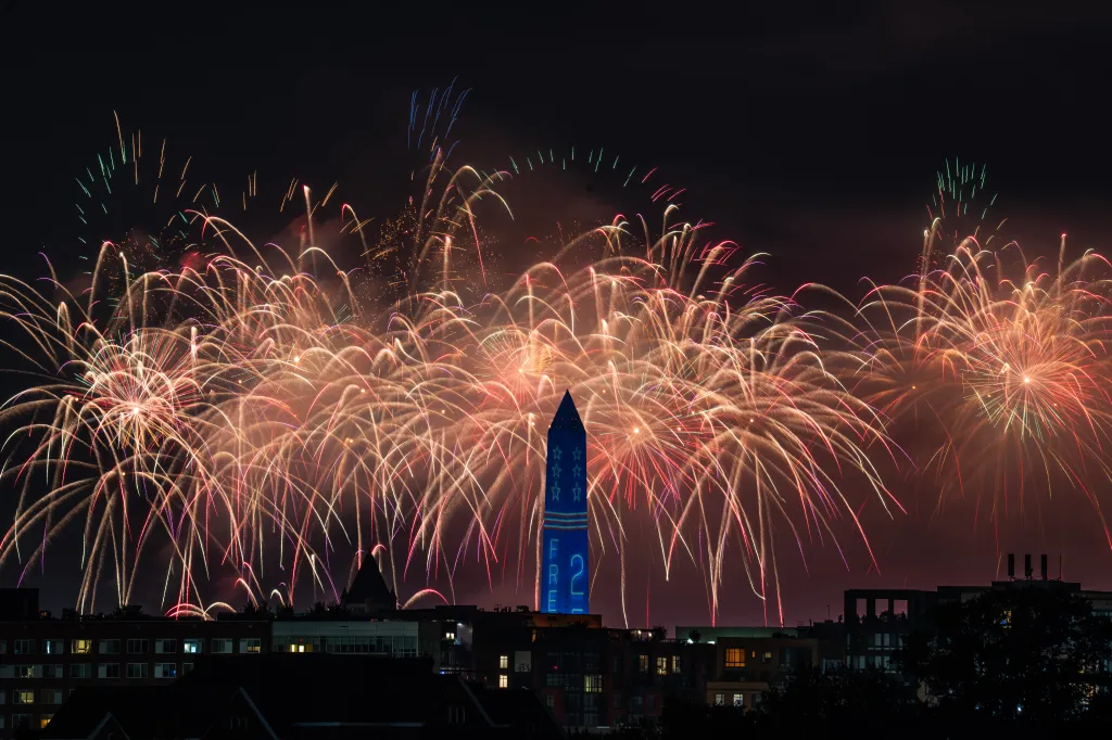 Fireworks bursting over a cityscape with a blue monument lit with stars and the text 