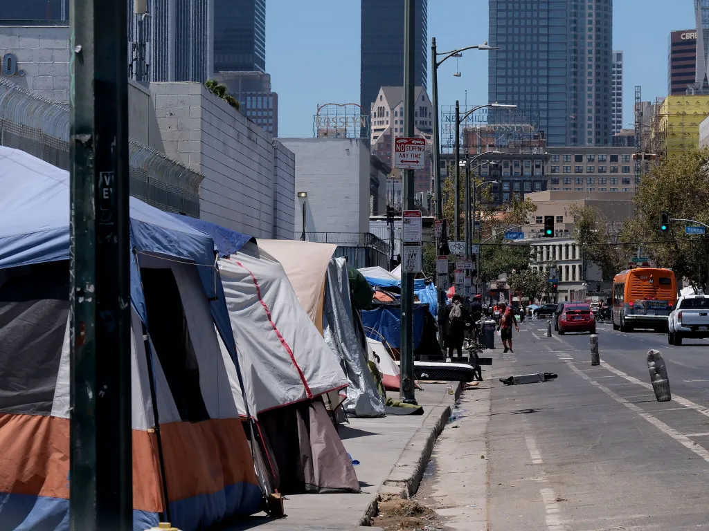 A row of tents on a sidewalk next to a street with cars, a bus, and people walking.